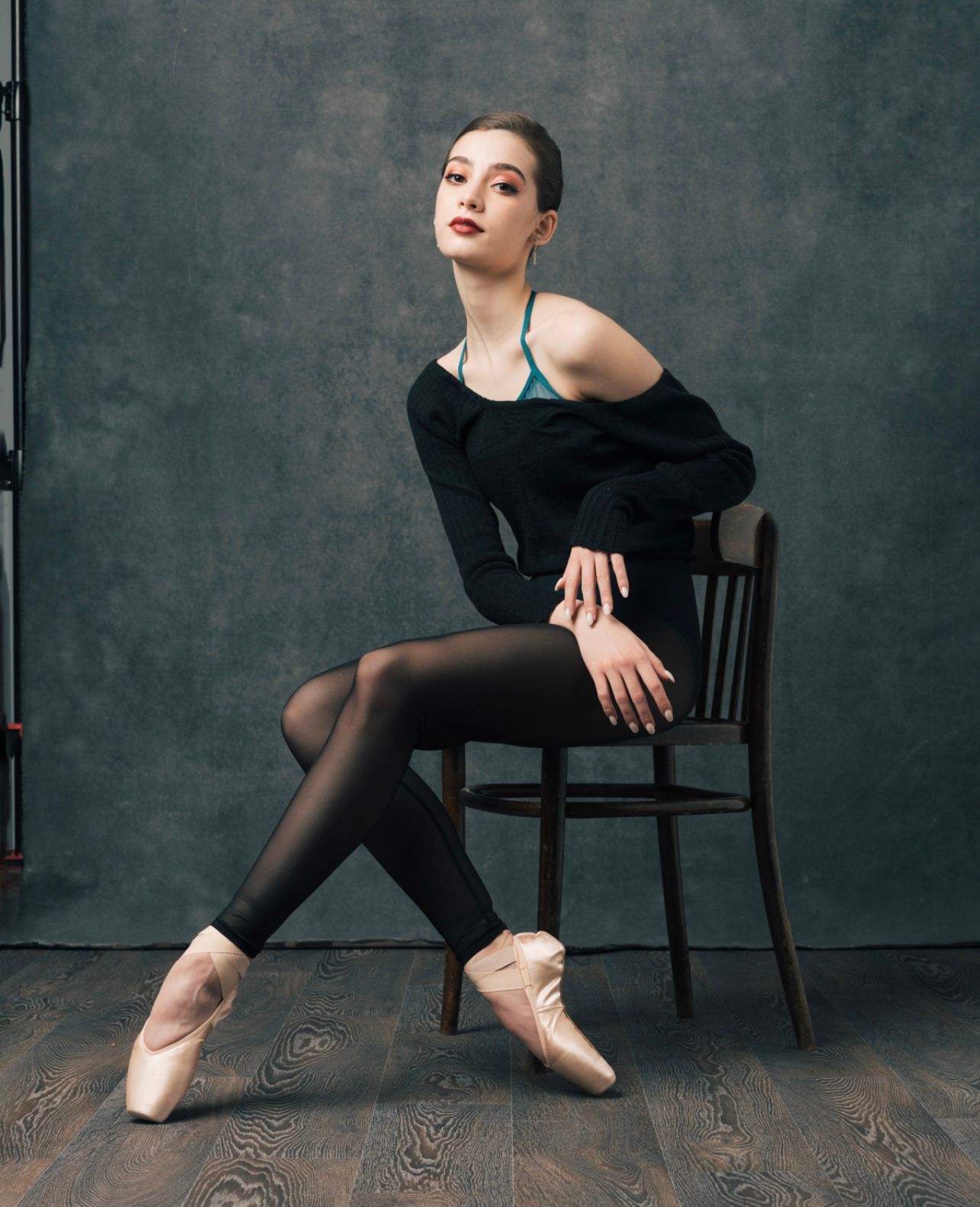 Ballet dancer seated, in pointe shoes, on a wooden chair; wearing a black sweater and tights; studio setting.