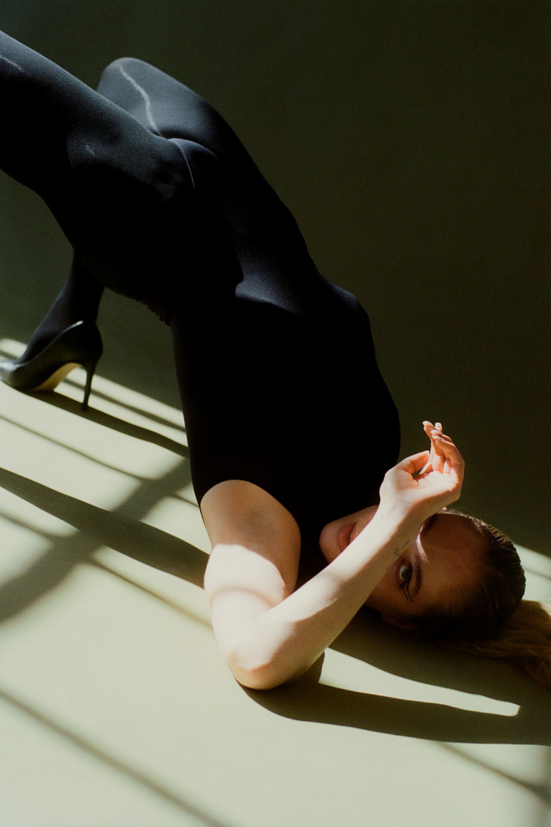 Woman in black leotard and heels, arched back, arm over face, lying on floor with sunlight shadows.