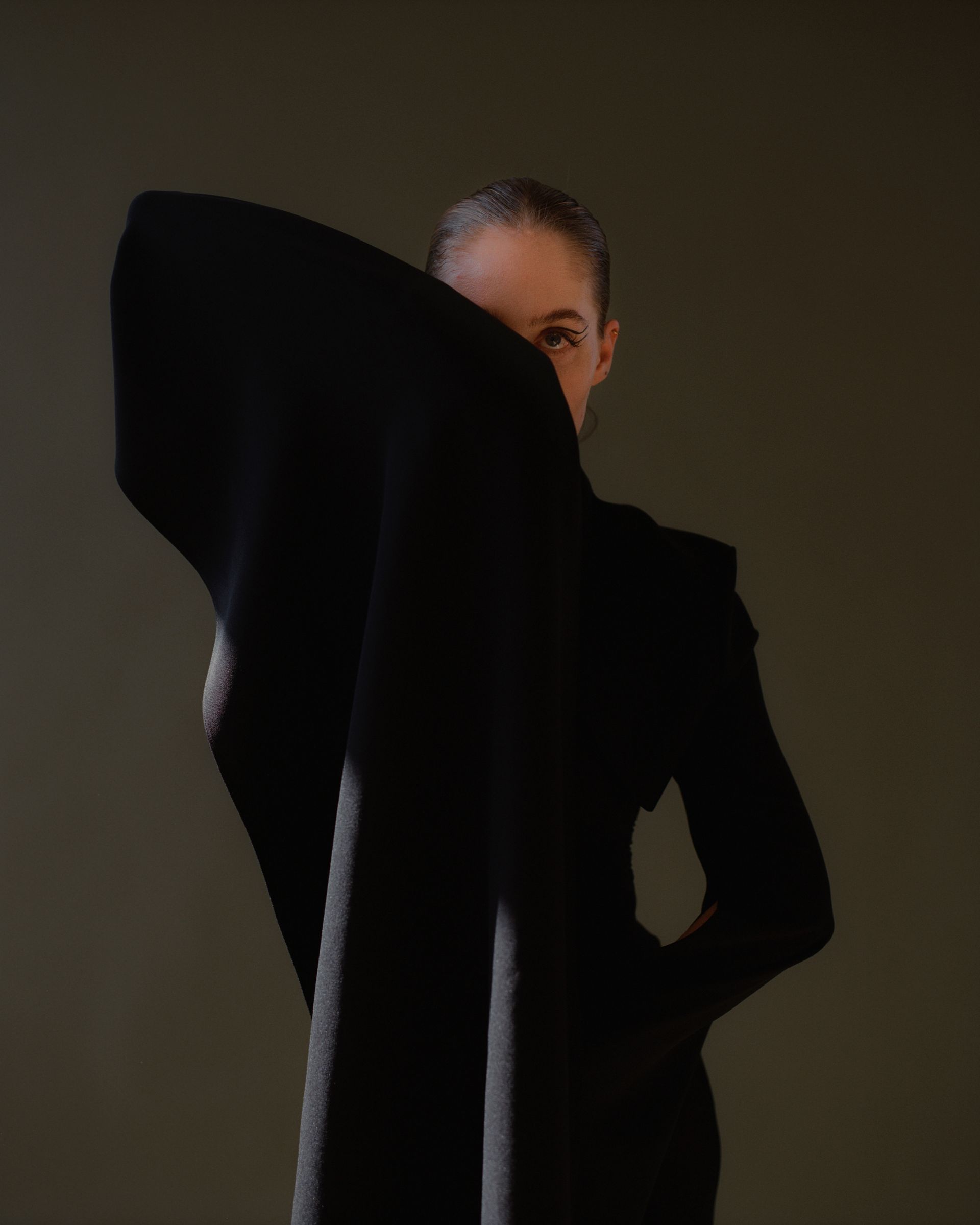 Woman in black dress, face partially covered by dark fabric, looking toward the viewer.