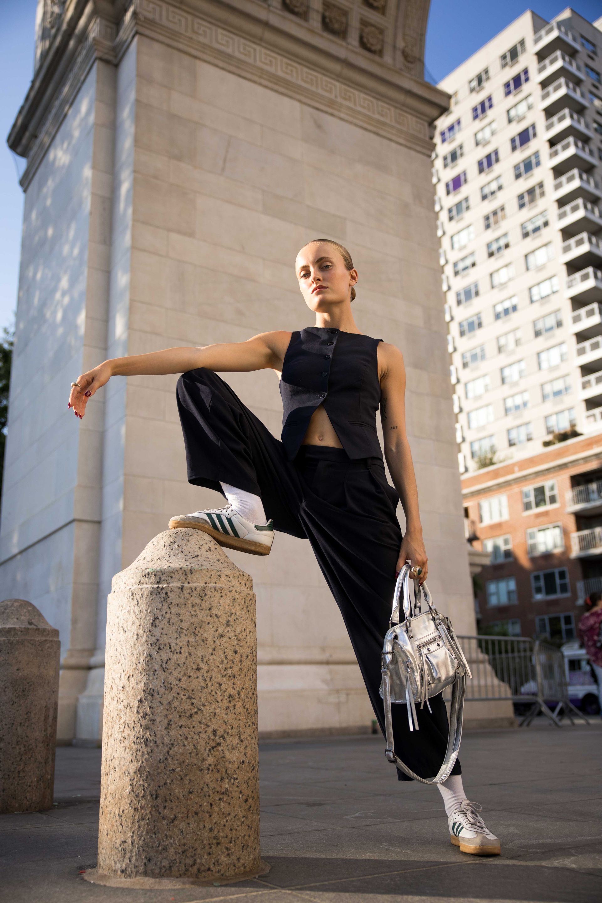 Woman in black outfit and white sneakers poses on a stone post, holding a silver bag near a large archway and building.