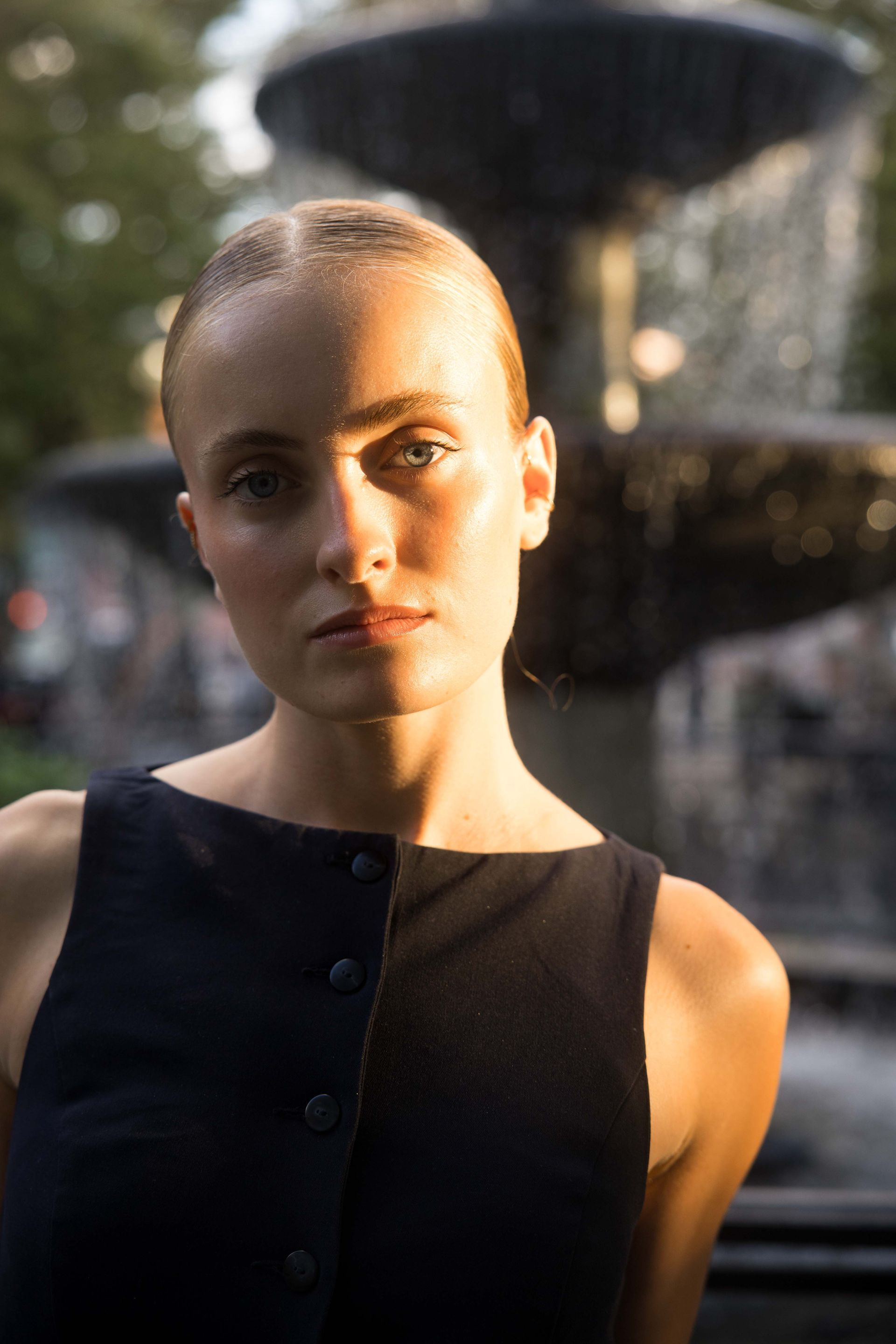 Woman in dark sleeveless top, lit by sunlight, in front of a fountain.