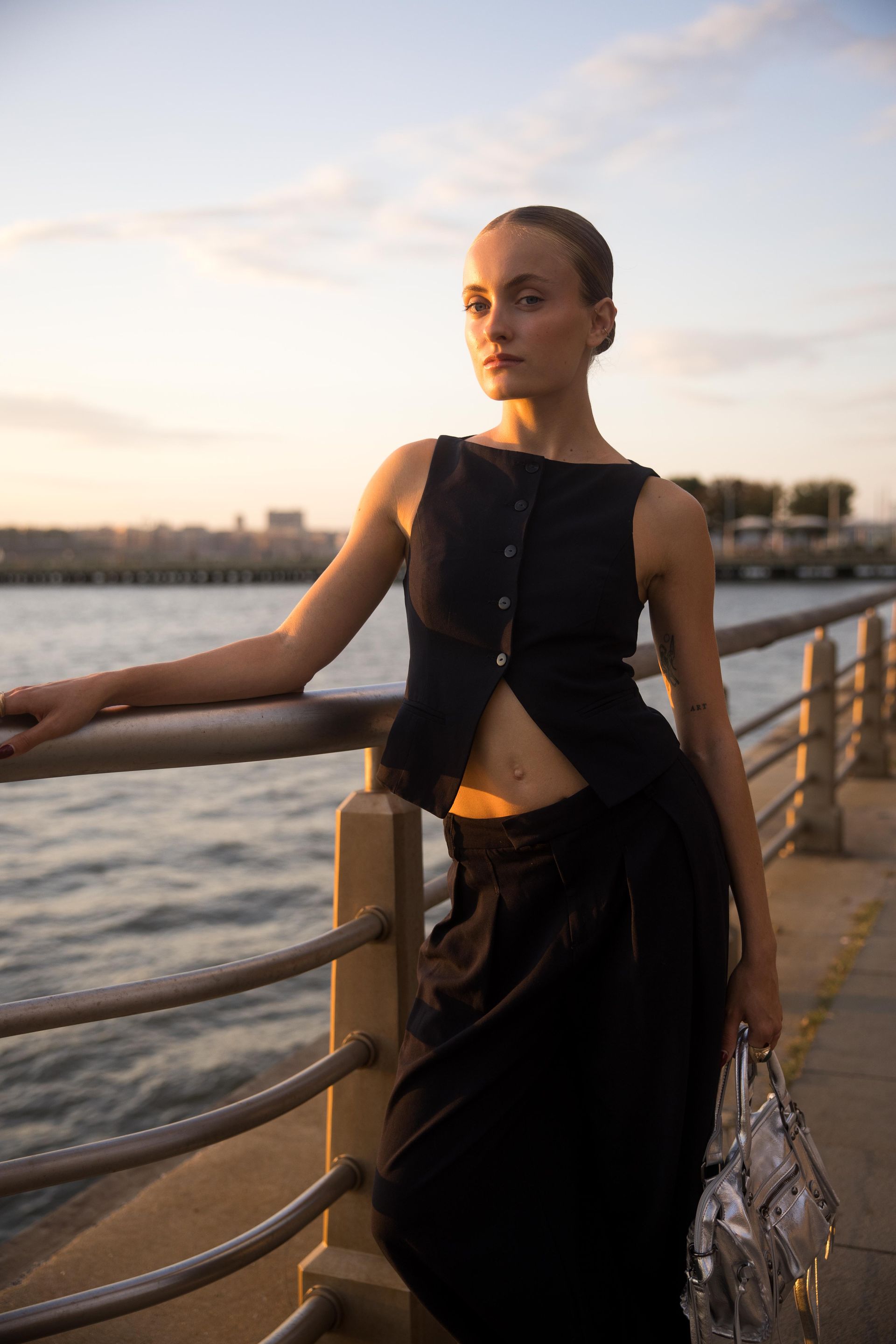 Woman in black outfit leans on railing by water at sunset.