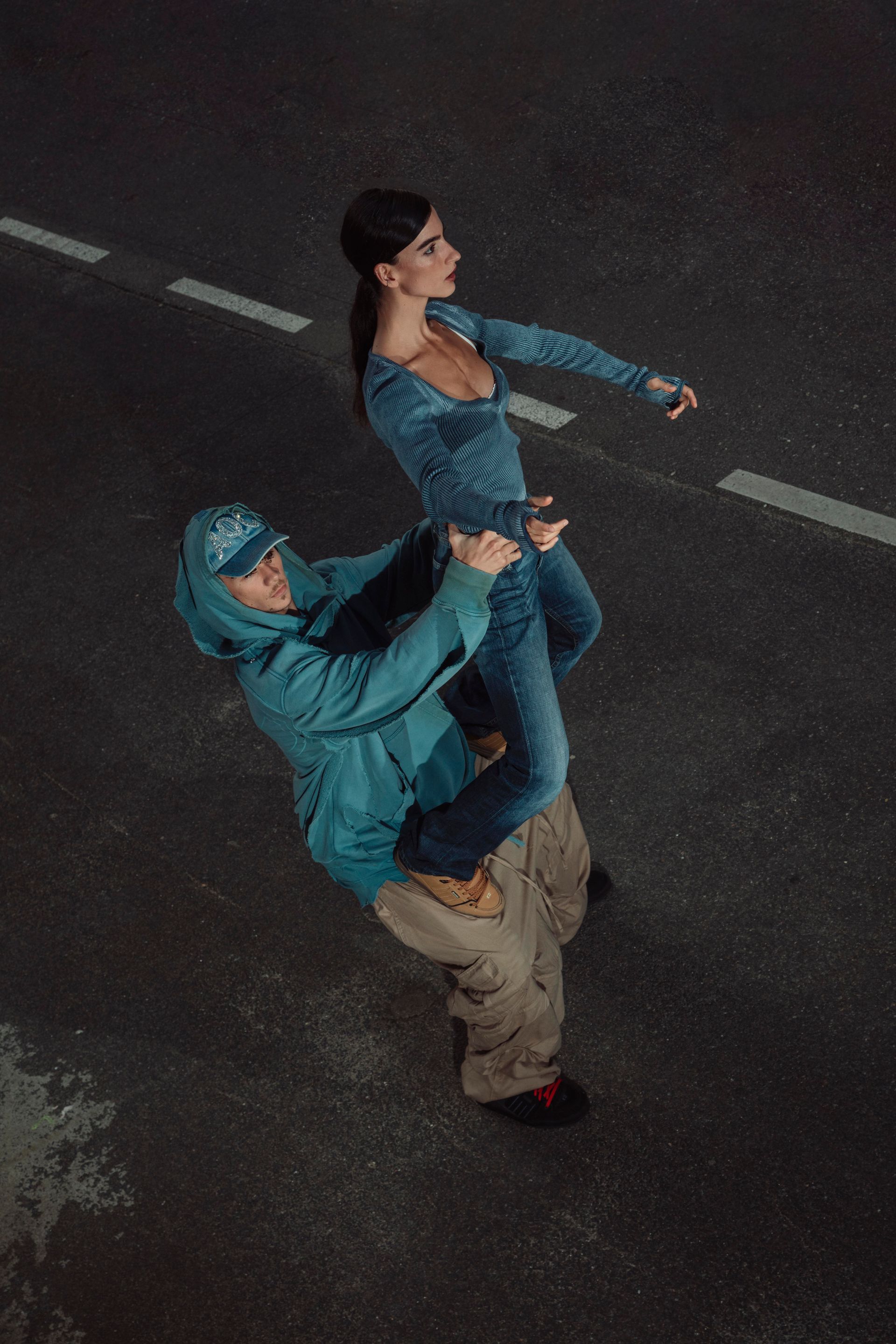 A person is carrying another person on their back on a road. They're wearing denim and a hoodie.