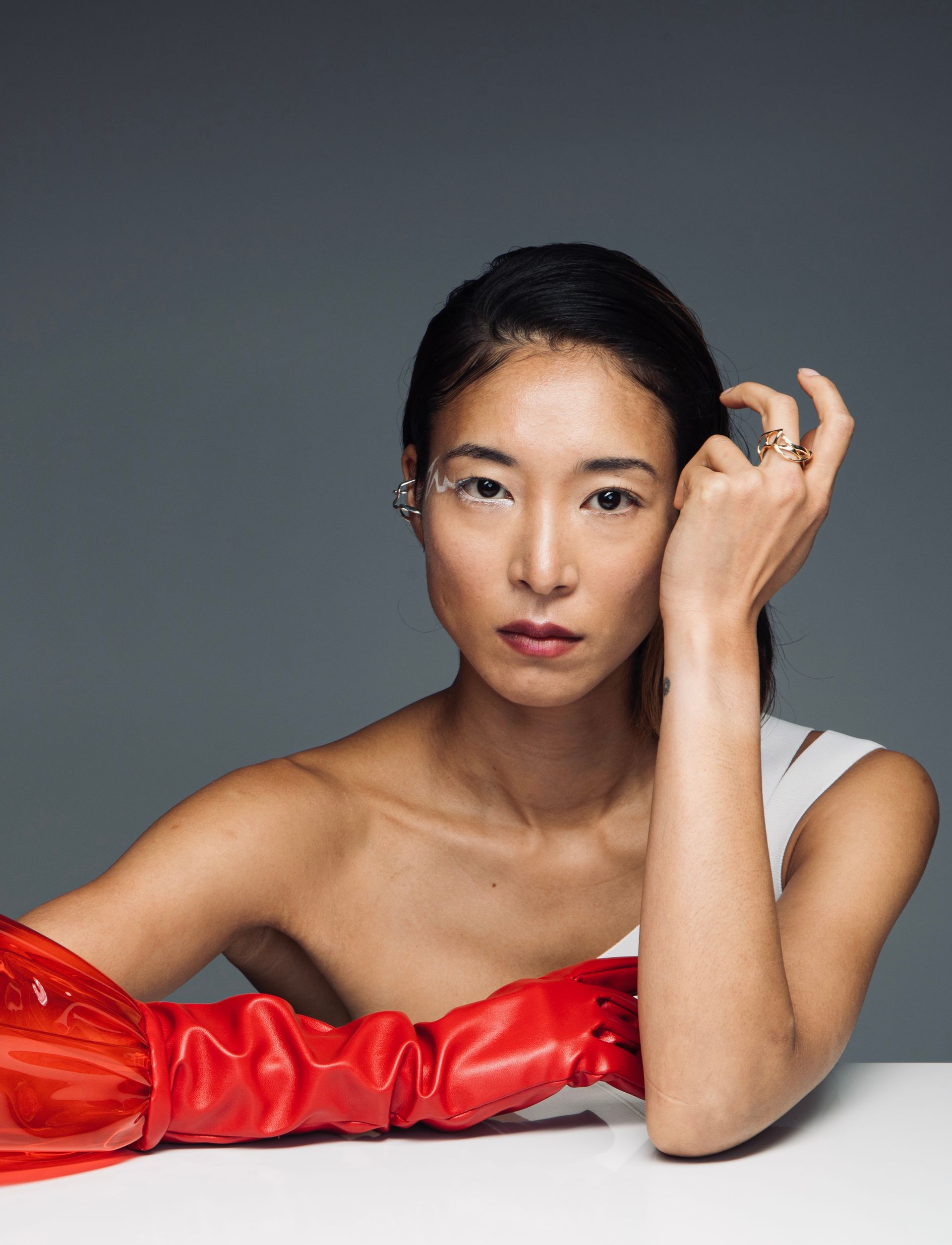 Woman with slicked-back hair, wearing a white top and a large red glove, leaning on a white surface with a gray background.