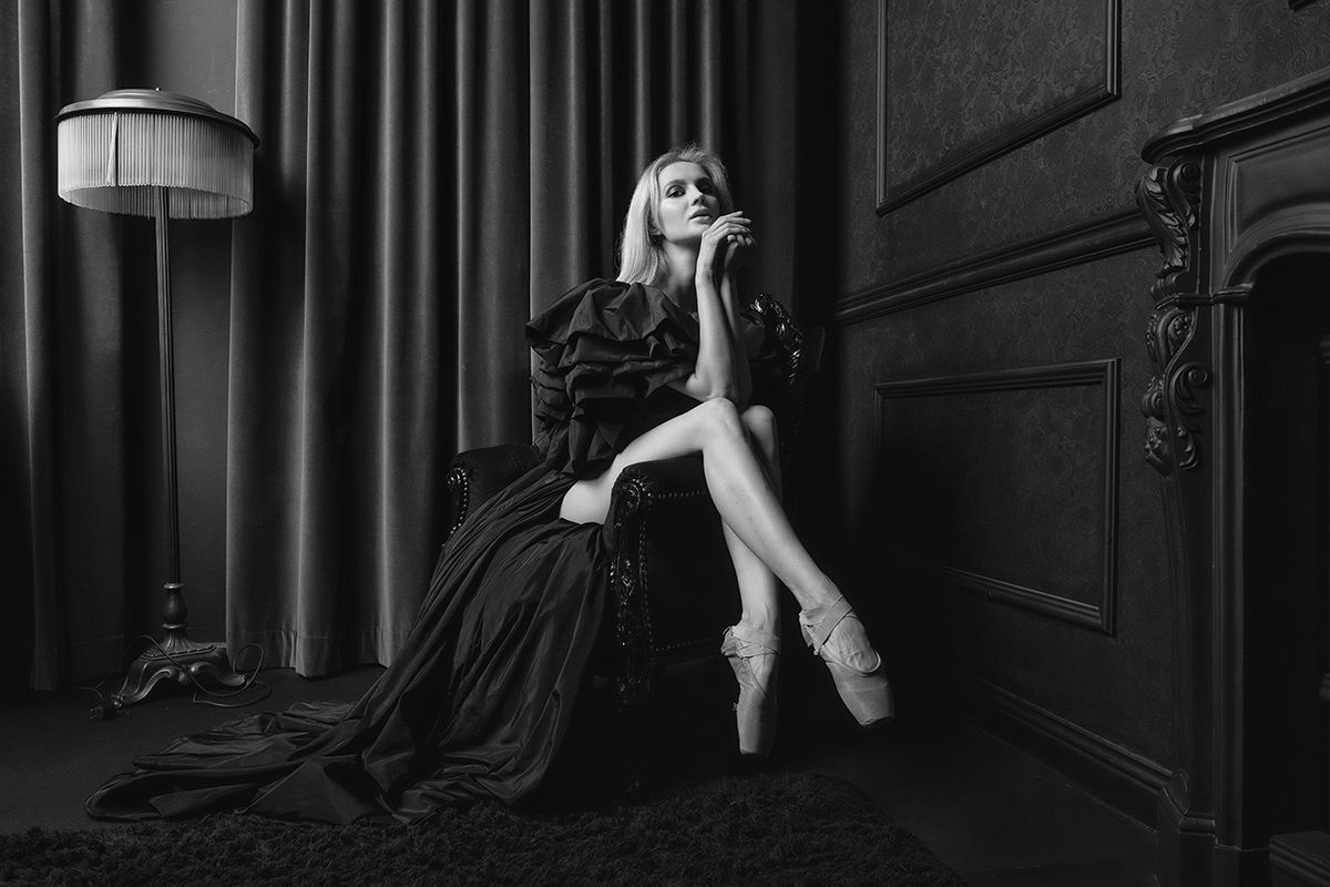 Woman in formal attire and pointe shoes, seated in a dimly lit room, posing with fingers near her face.