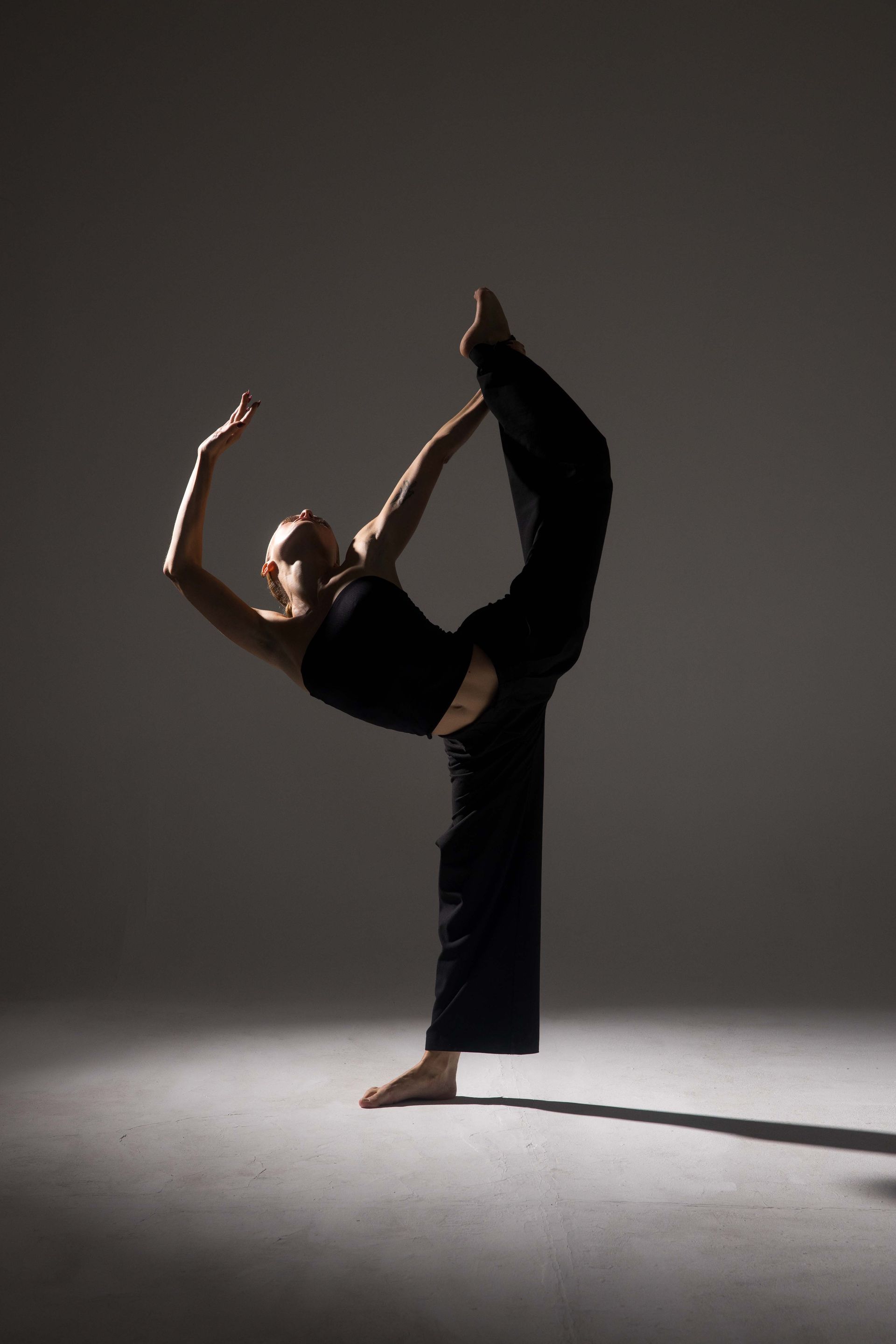 Dancer in black outfit balances, arching back, lifting leg overhead. Studio lit with spotlight, shadow below.