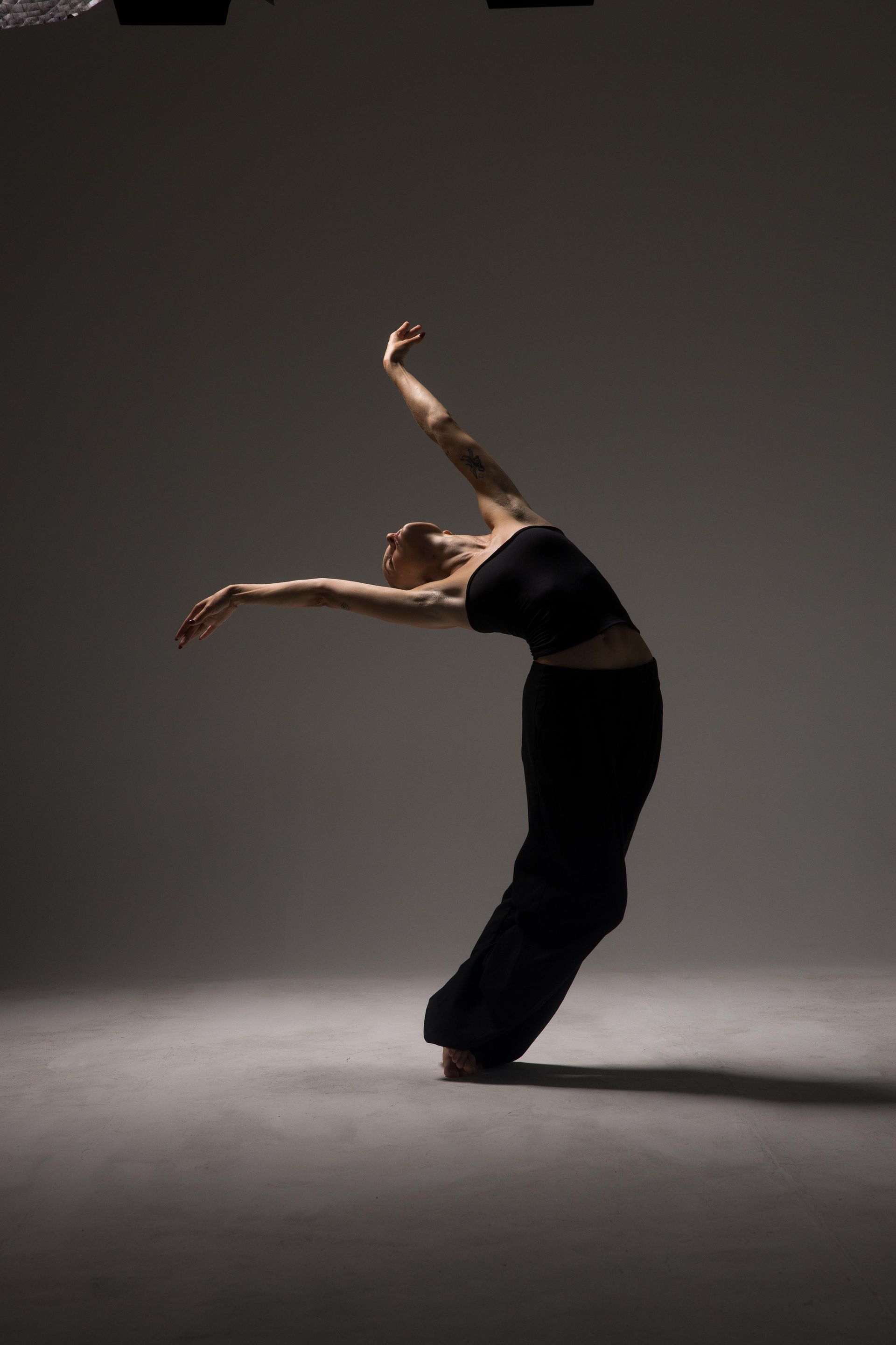 Dancer in black outfit arches back, arms extended, in a studio with spotlight.