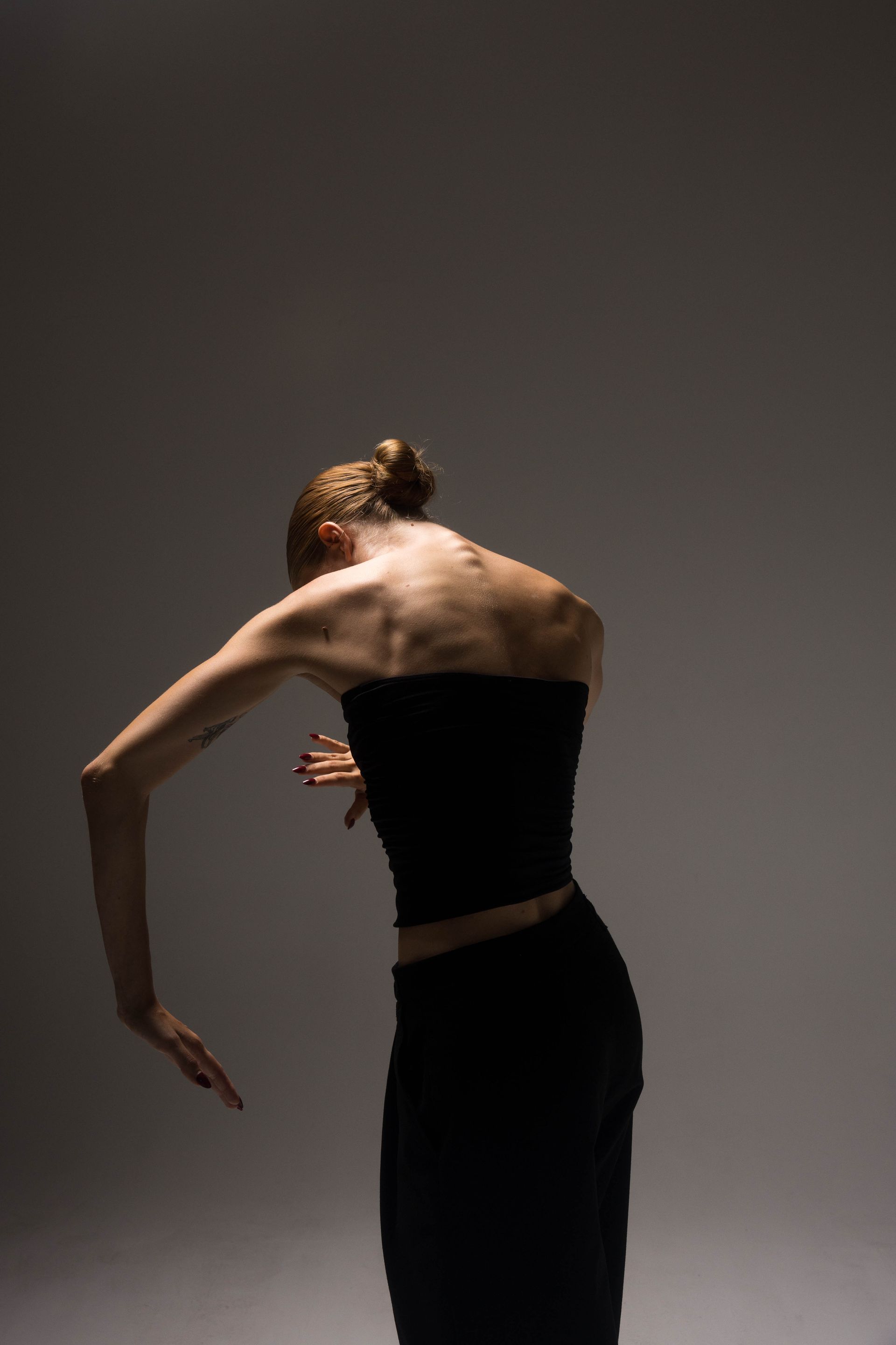 Woman in black clothing, arched back, arms extended, lit from above against a gray backdrop.