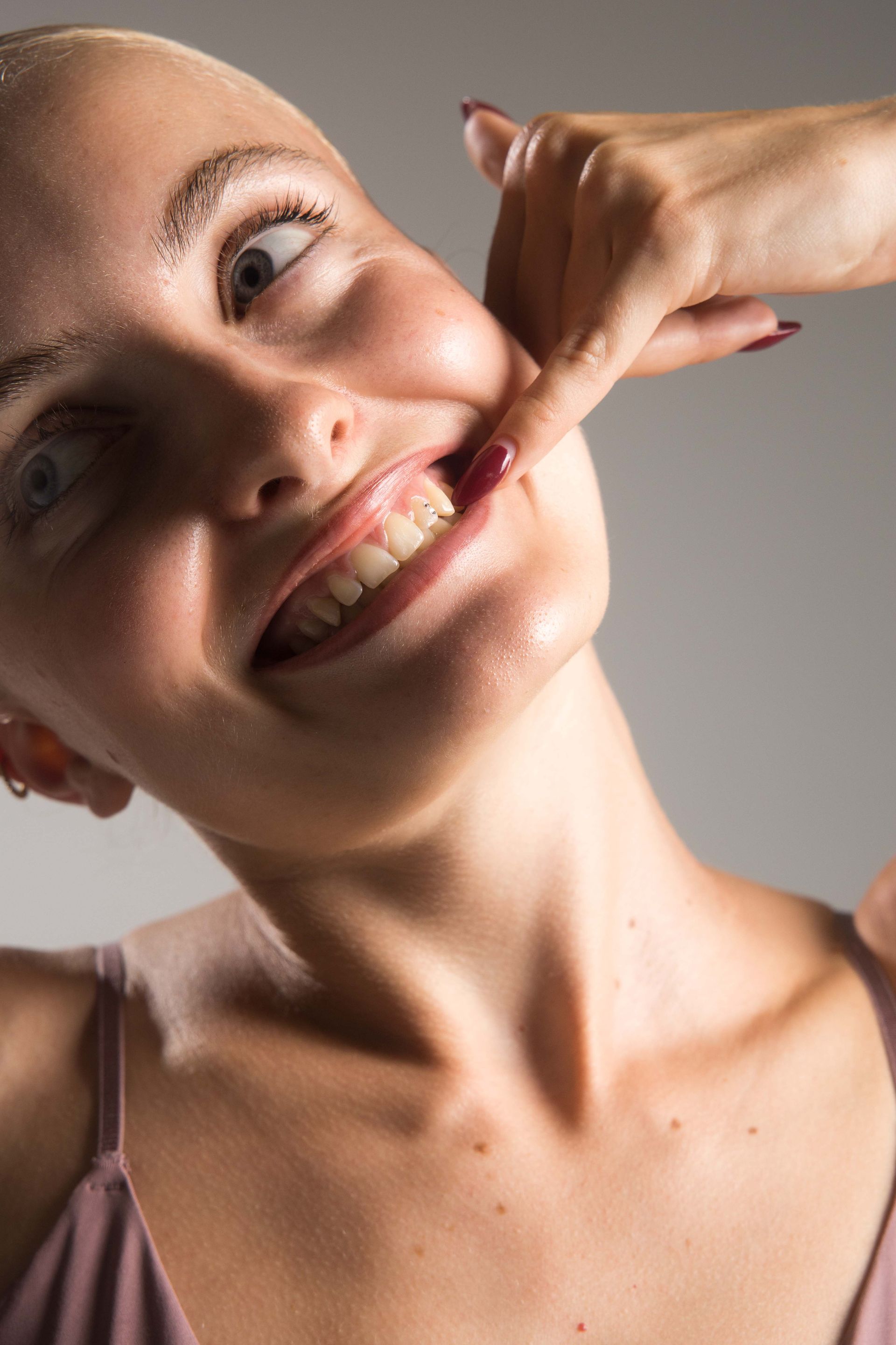 Woman's face pulled, showing teeth, by another hand, pale background.