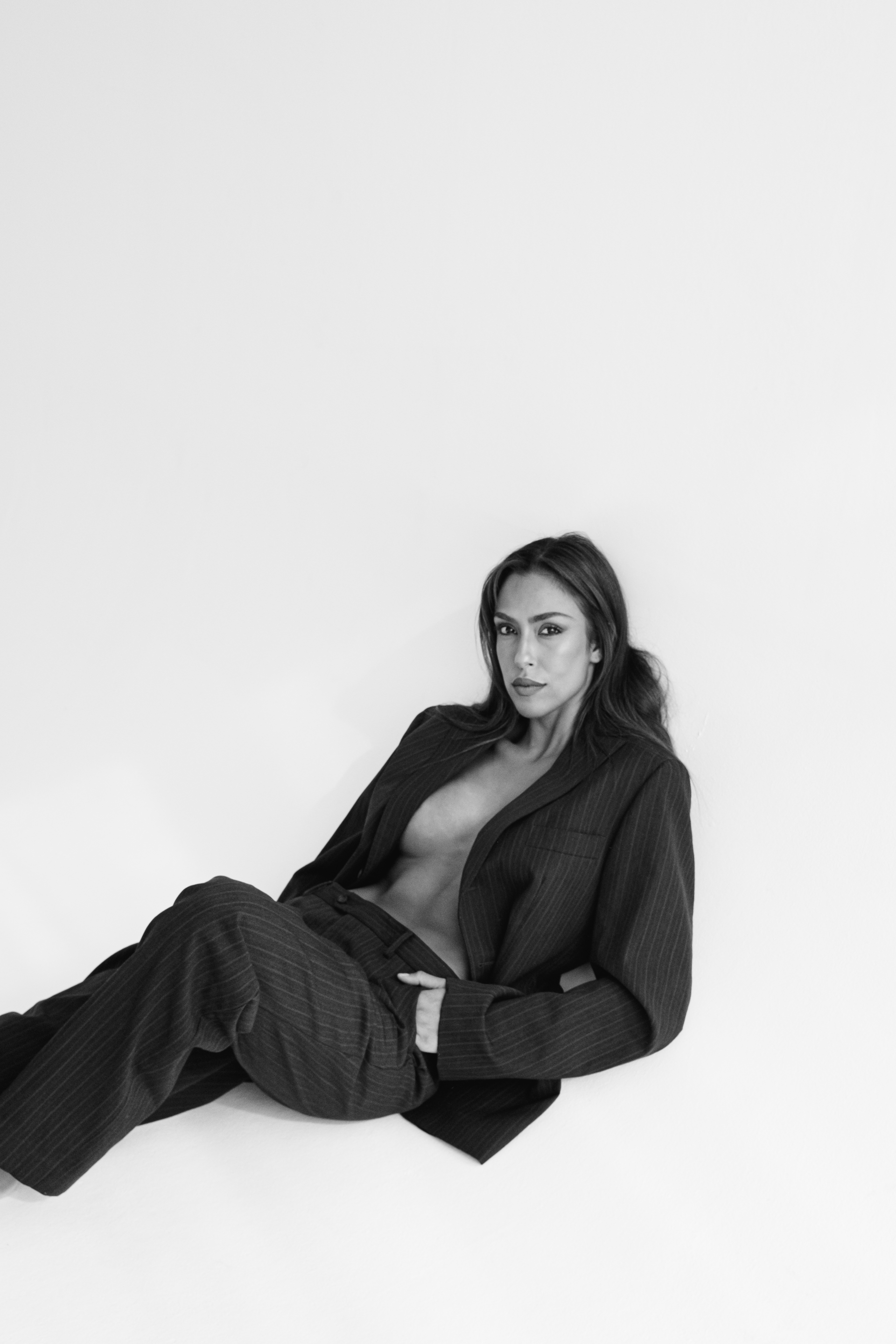 Woman in a dark pinstripe suit, leaning against a white background with one hand in her pocket.