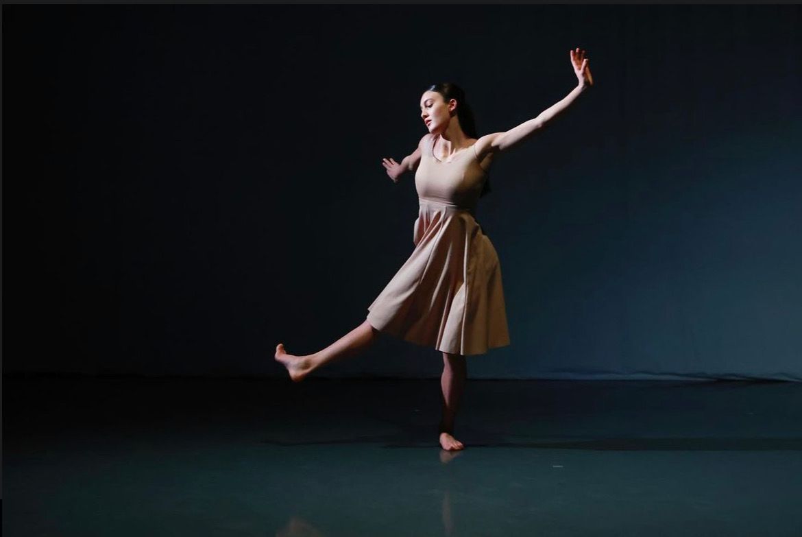 Dancer in a beige dress on a dark stage, arms extended, leg lifted.