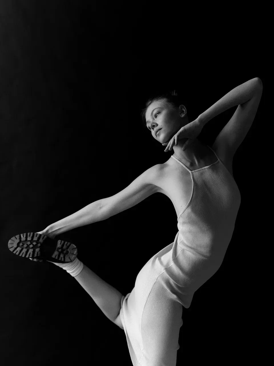 Woman in a white dress and sock, balancing with one leg extended, arm raised; black background.