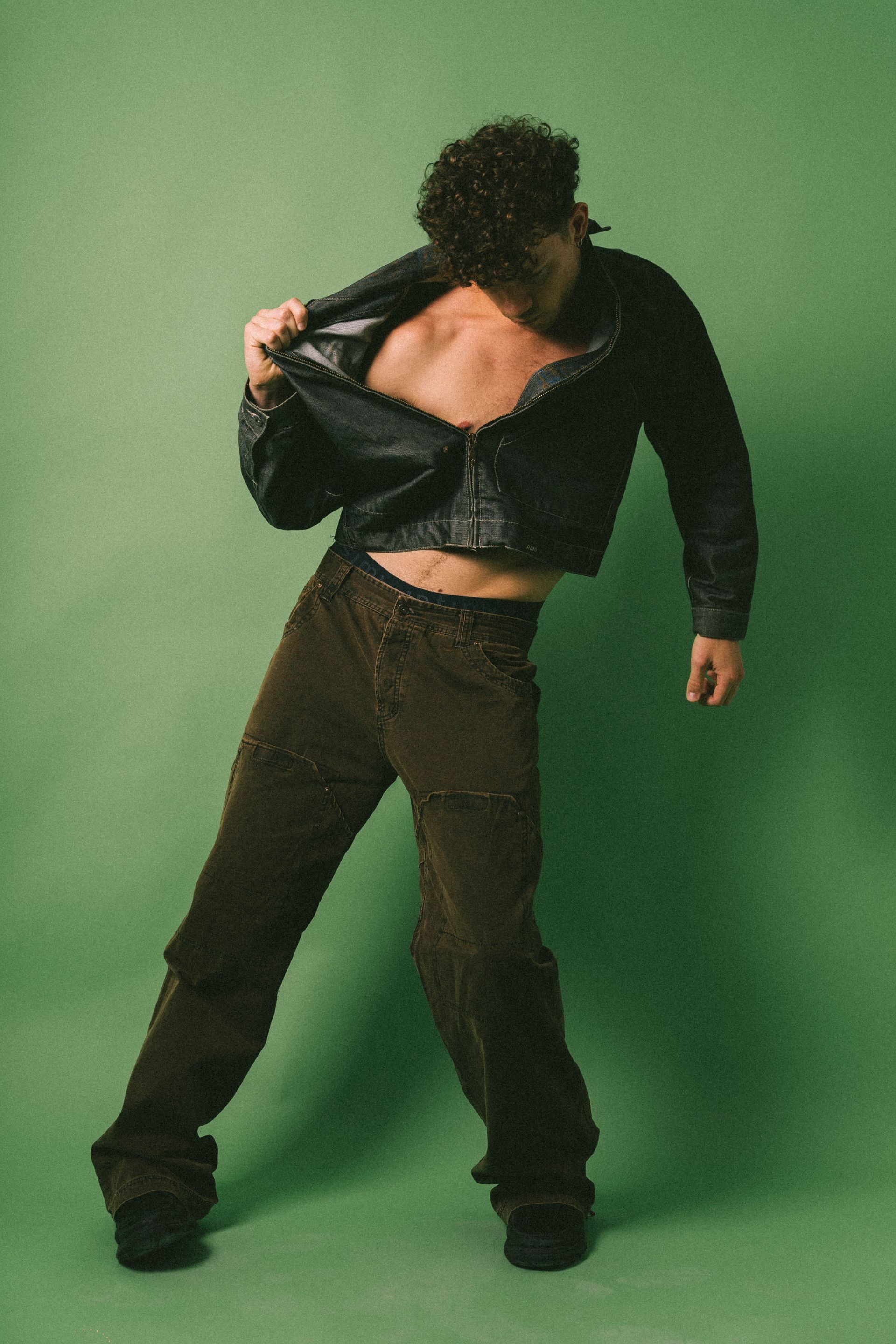 Man in black leather jacket and brown pants, pulling open jacket, torso exposed, green background.