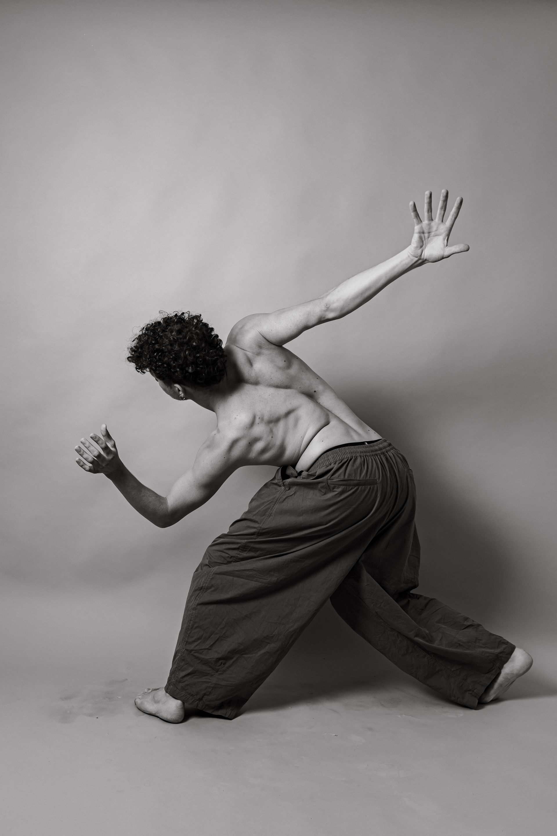 Man in loose pants, dancing; one arm extended, other bent, back arched, grayscale.
