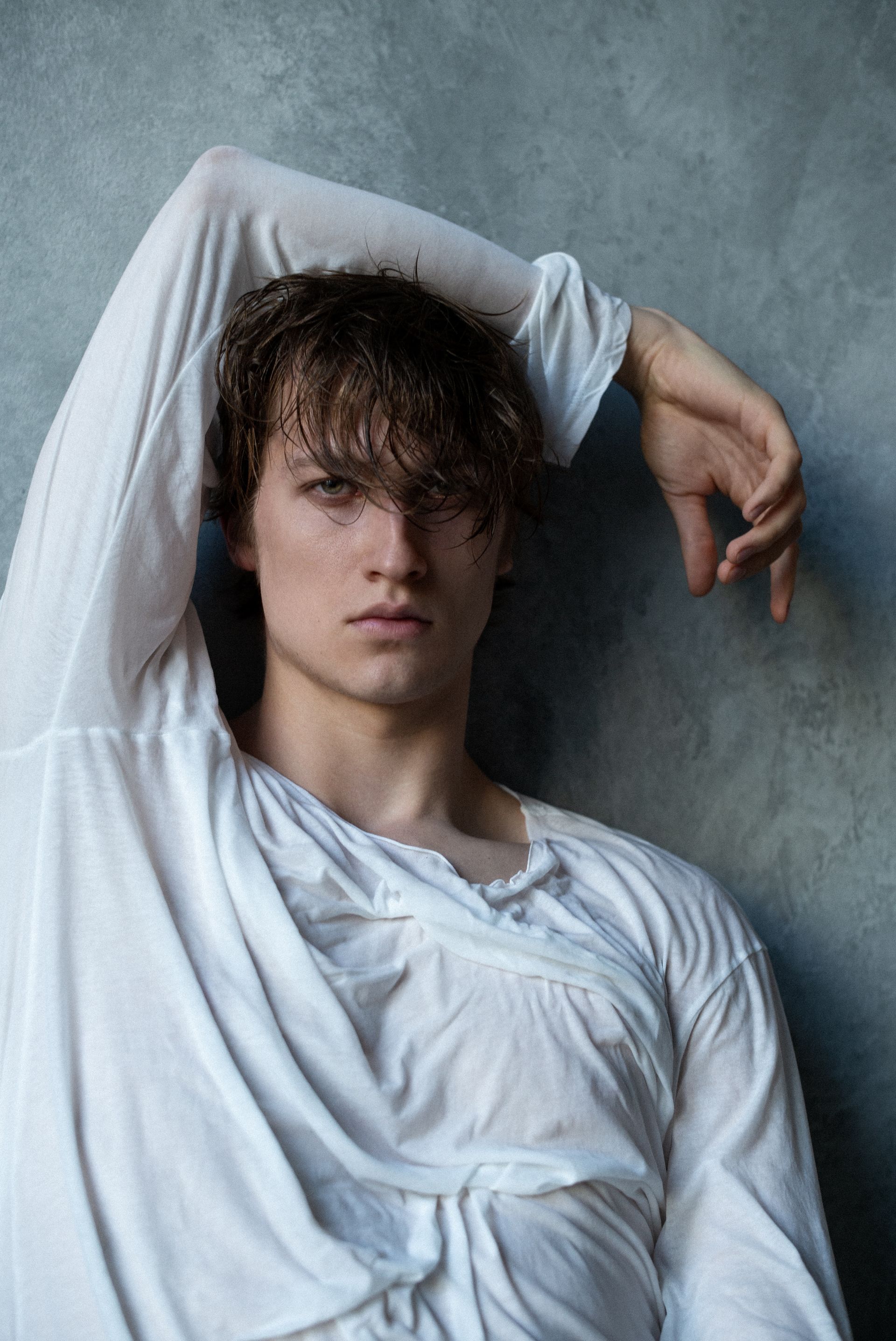Man in white shirt leans against a blue-gray wall, arm raised. His gaze is direct and serious.