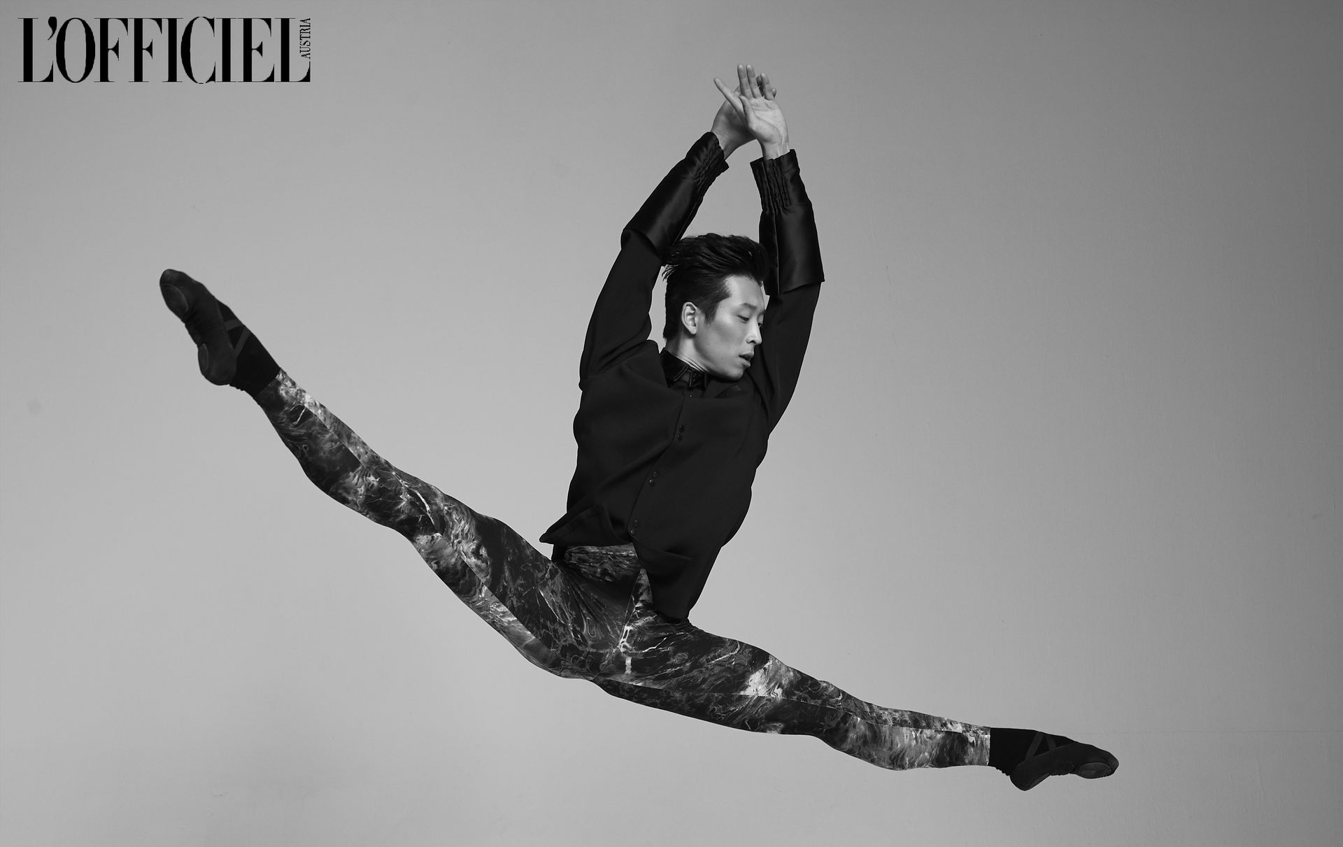 A dancer in black shirt and sparkly pants leaps in a split, arms overhead.