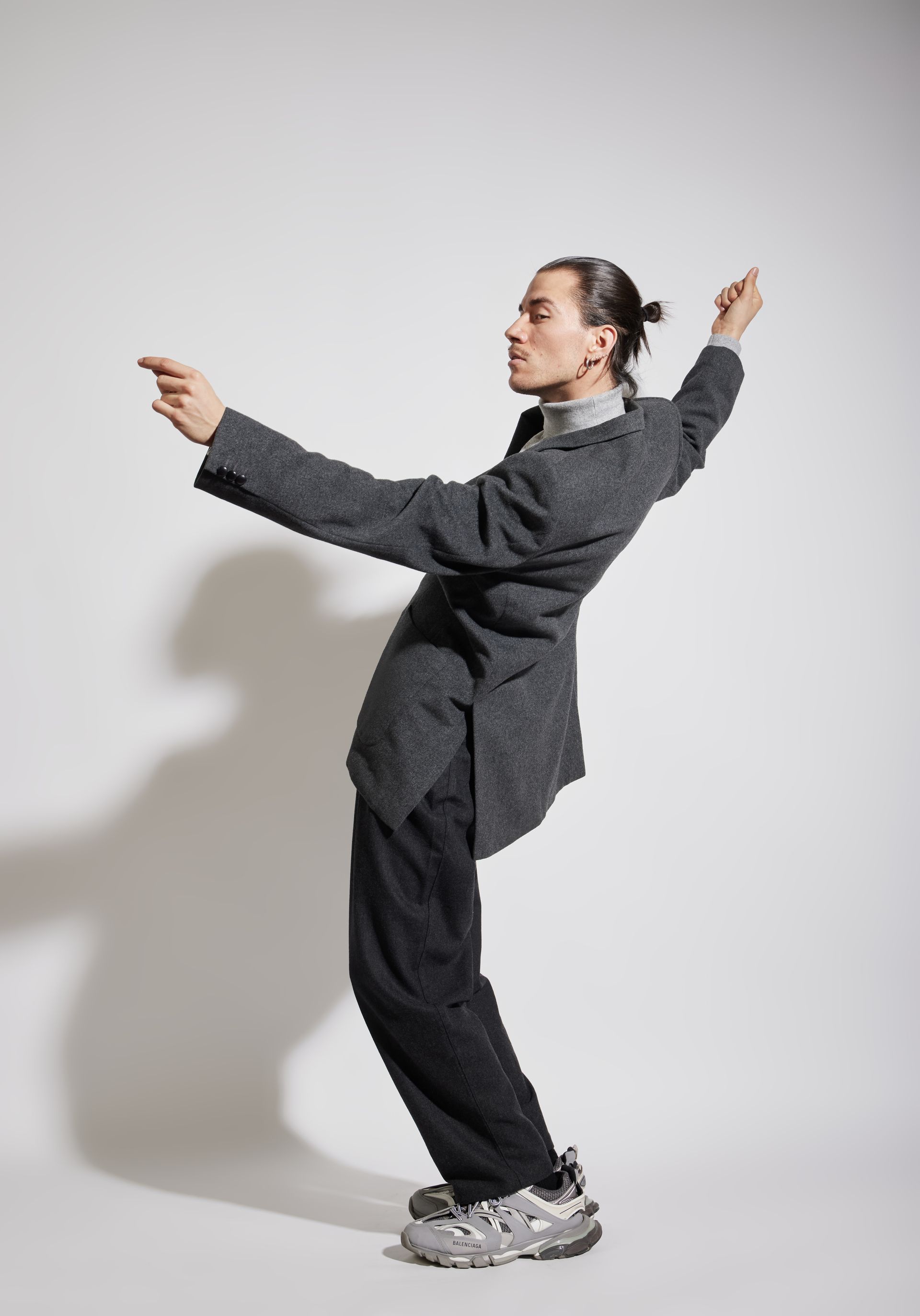 Man in gray suit and sneakers, leaning back with arms outstretched, against a white background.