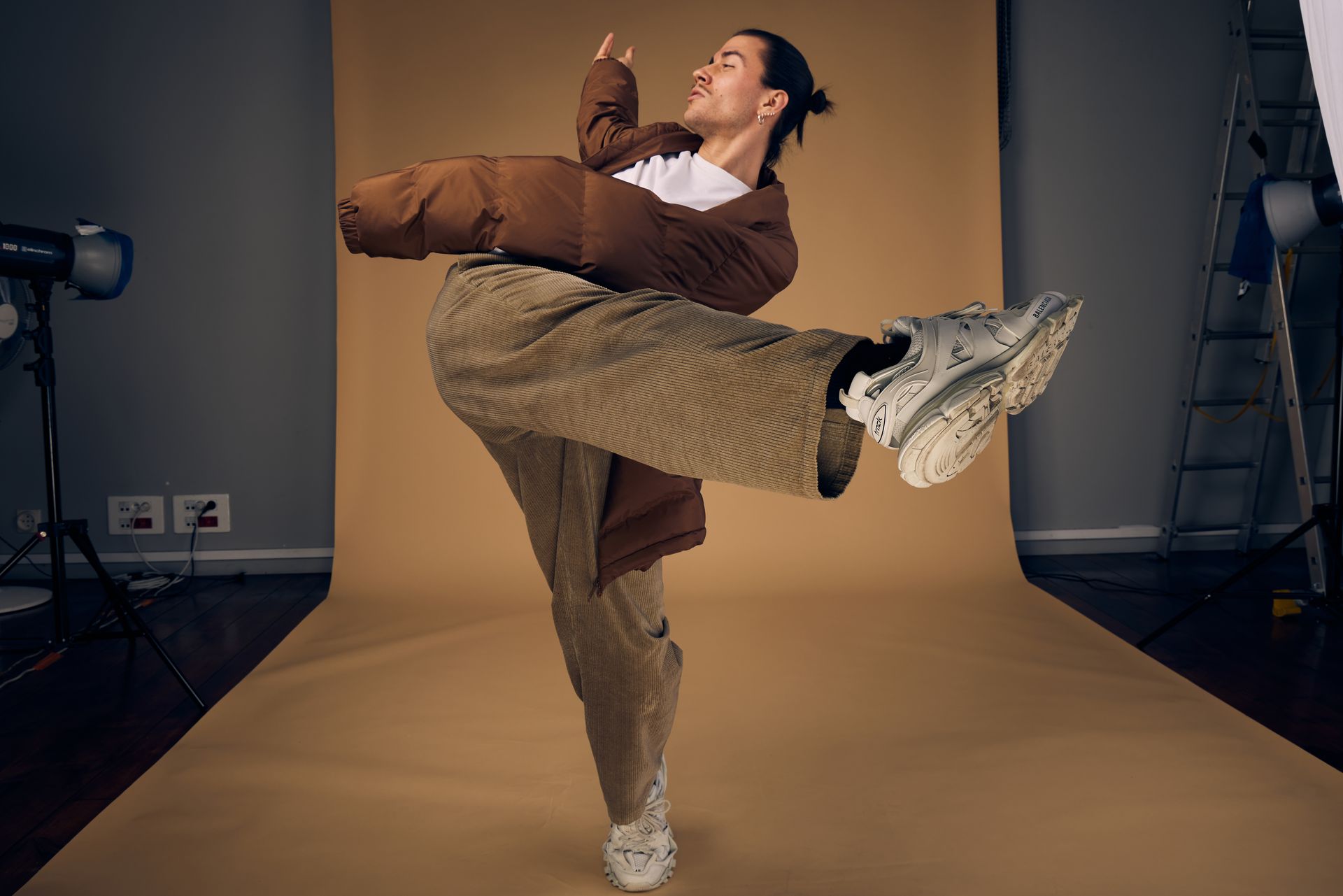 Person in brown jacket and pants poses with a leg raised in a studio setting with a tan backdrop.