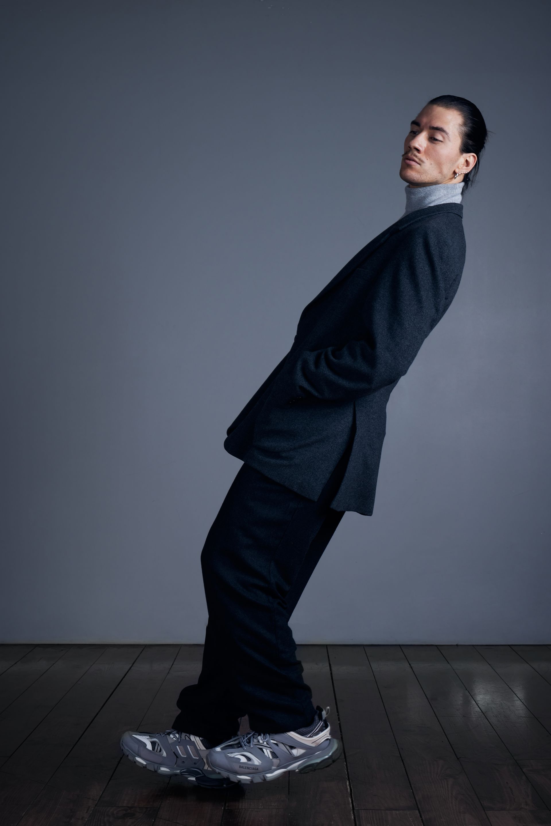 Man in suit and sneakers leaning back on one leg. Wearing a gray turtleneck. Posing in front of a gray wall.