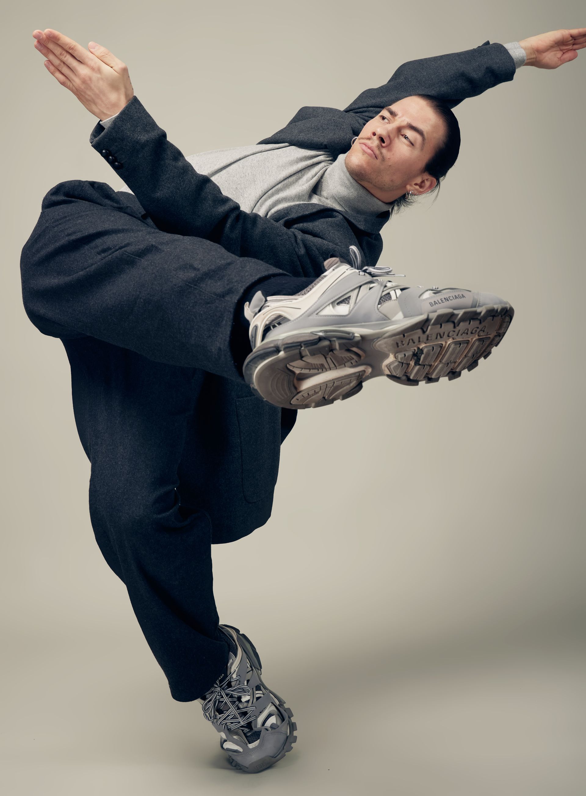 Person in suit and sneakers, mid-air pose. Gray suit, light turtleneck, gray sneakers, light background.