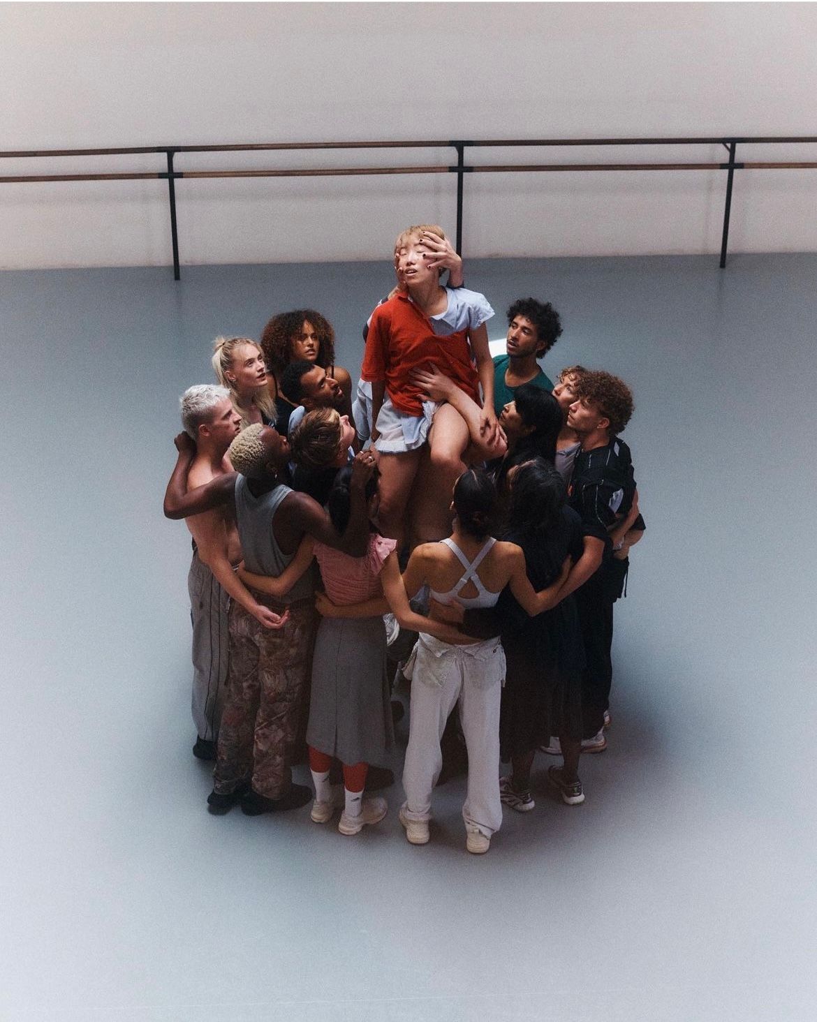 A group of people embrace, one person is seated atop them. They are in a dance studio with a barre.