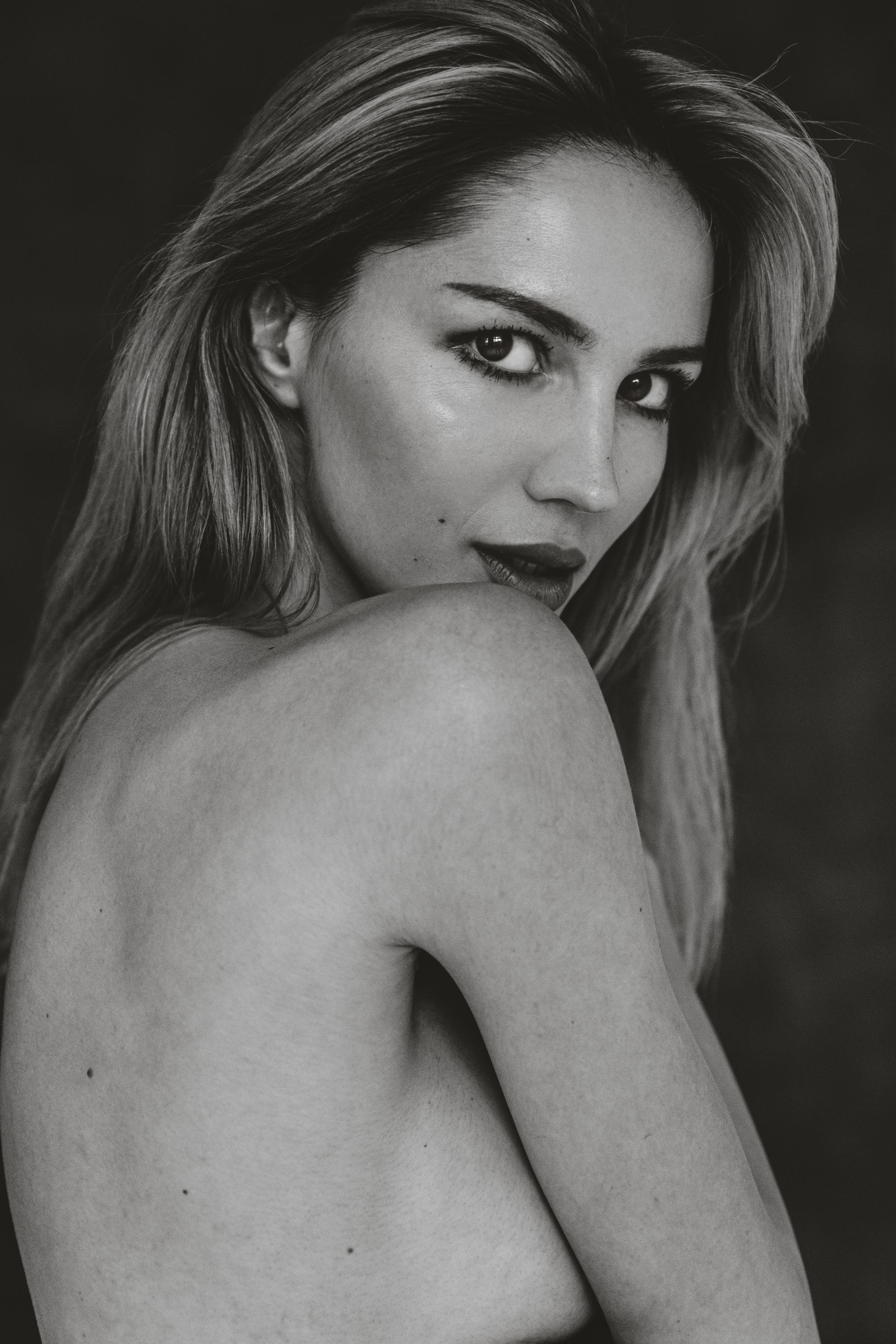 Woman with long blonde hair looks over shoulder, dark eye makeup, partially nude, black and white.