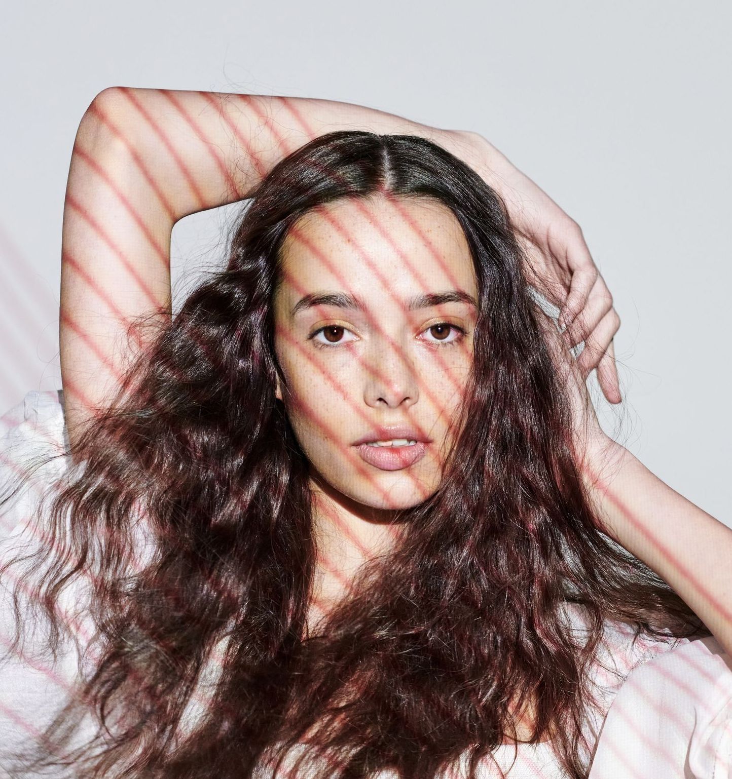 Woman with long, wavy brown hair, illuminated by striped shadows, looking towards the viewer.