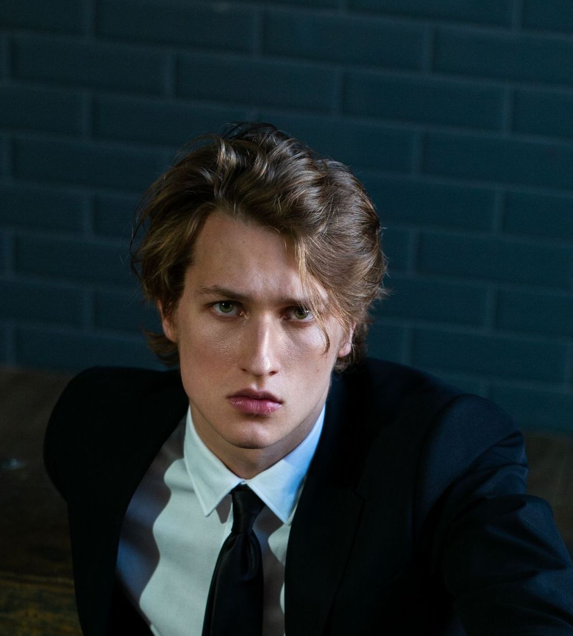 Man in black suit, white shirt, and black tie, looking directly at the viewer against a blue brick wall.