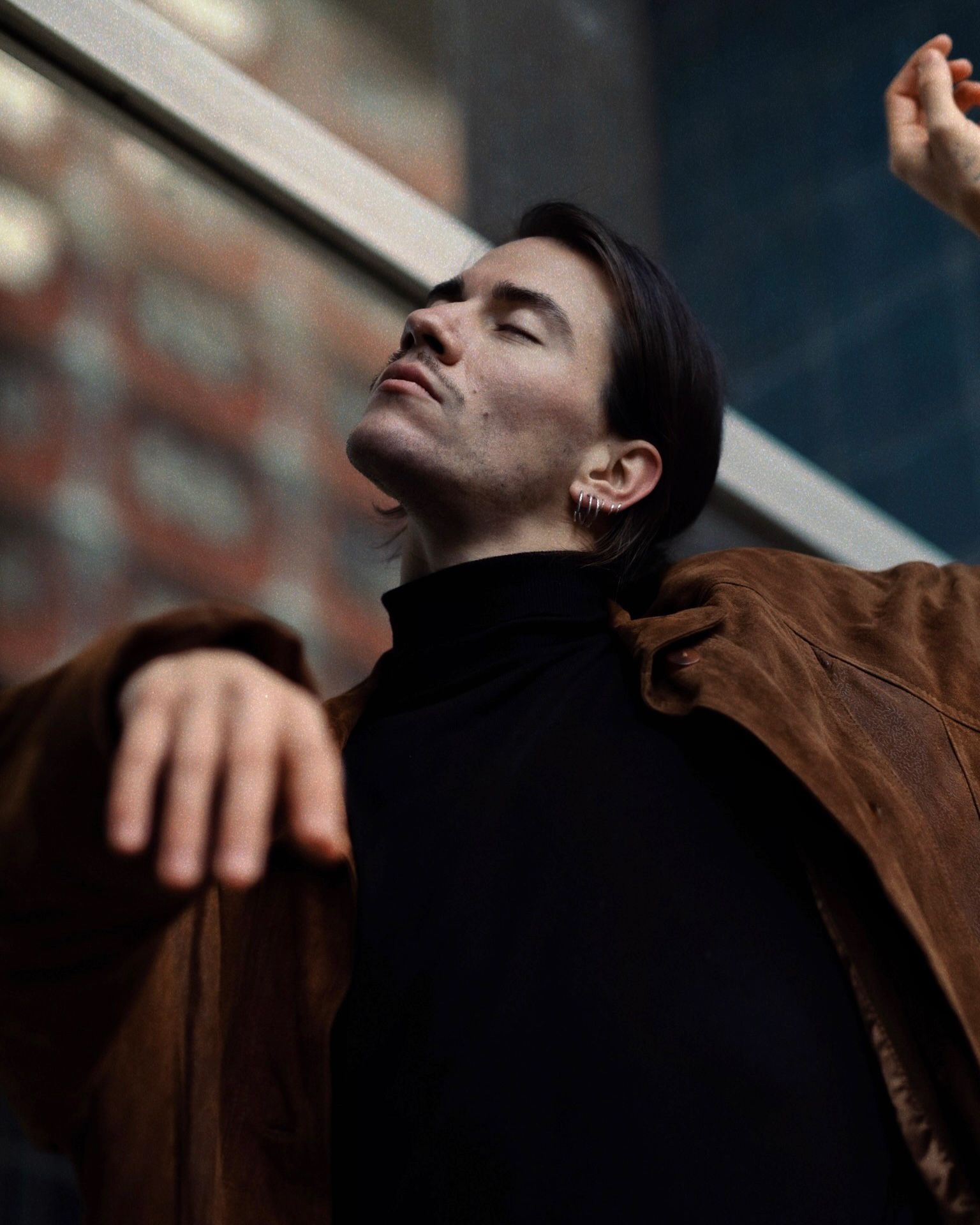 Man with eyes closed, arms outstretched, wearing a black turtleneck and brown jacket, in front of a building.