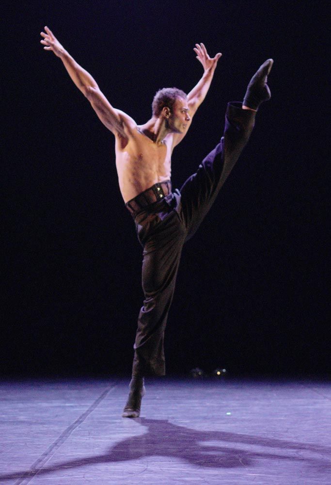 Male dancer in mid-air, leg extended upward. Arms raised, torso bare, wearing black pants, on stage.