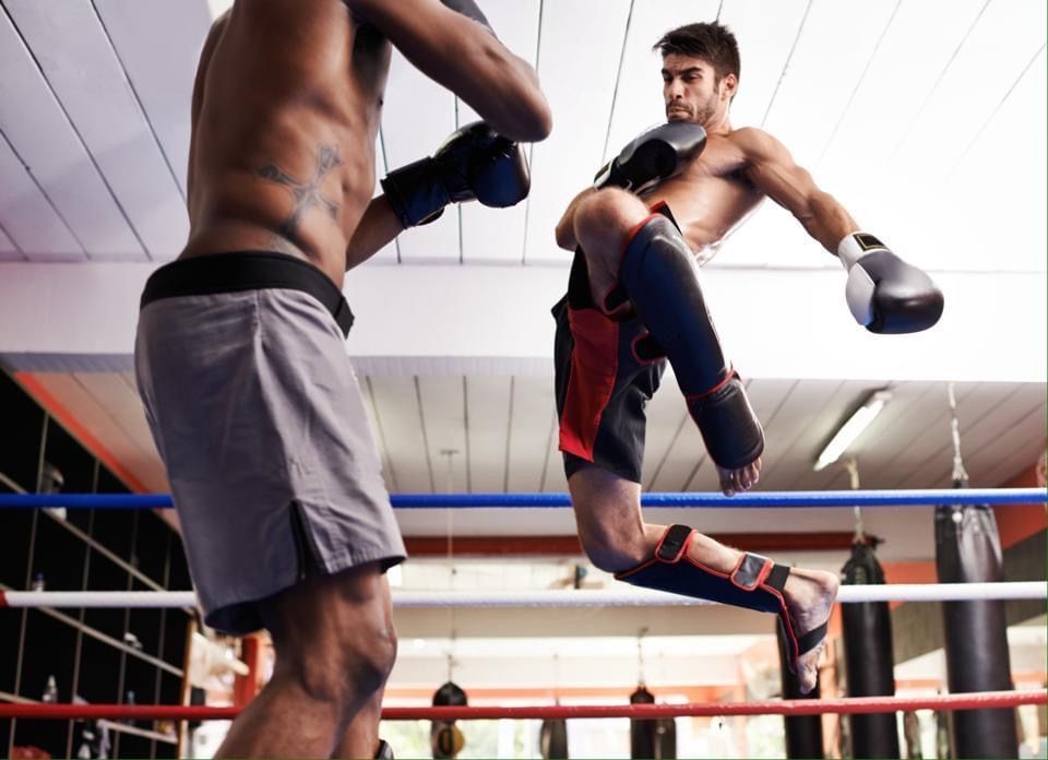 Two men in a boxing ring; one kicks, other blocks. Both wear gloves and shorts. Gym setting.