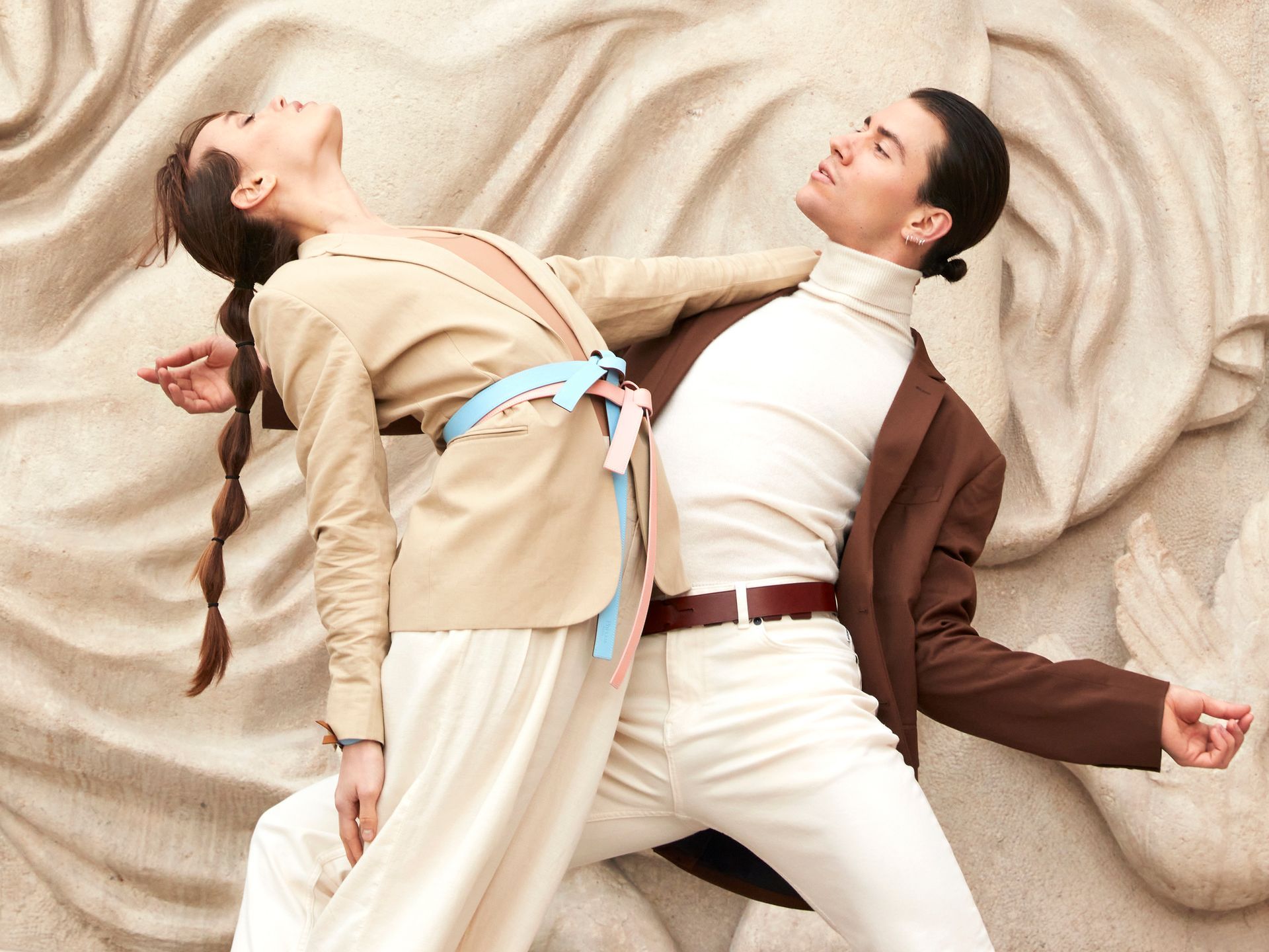 Couple in stylish clothing leaning against a stone wall, arms outstretched, with neutral expressions.