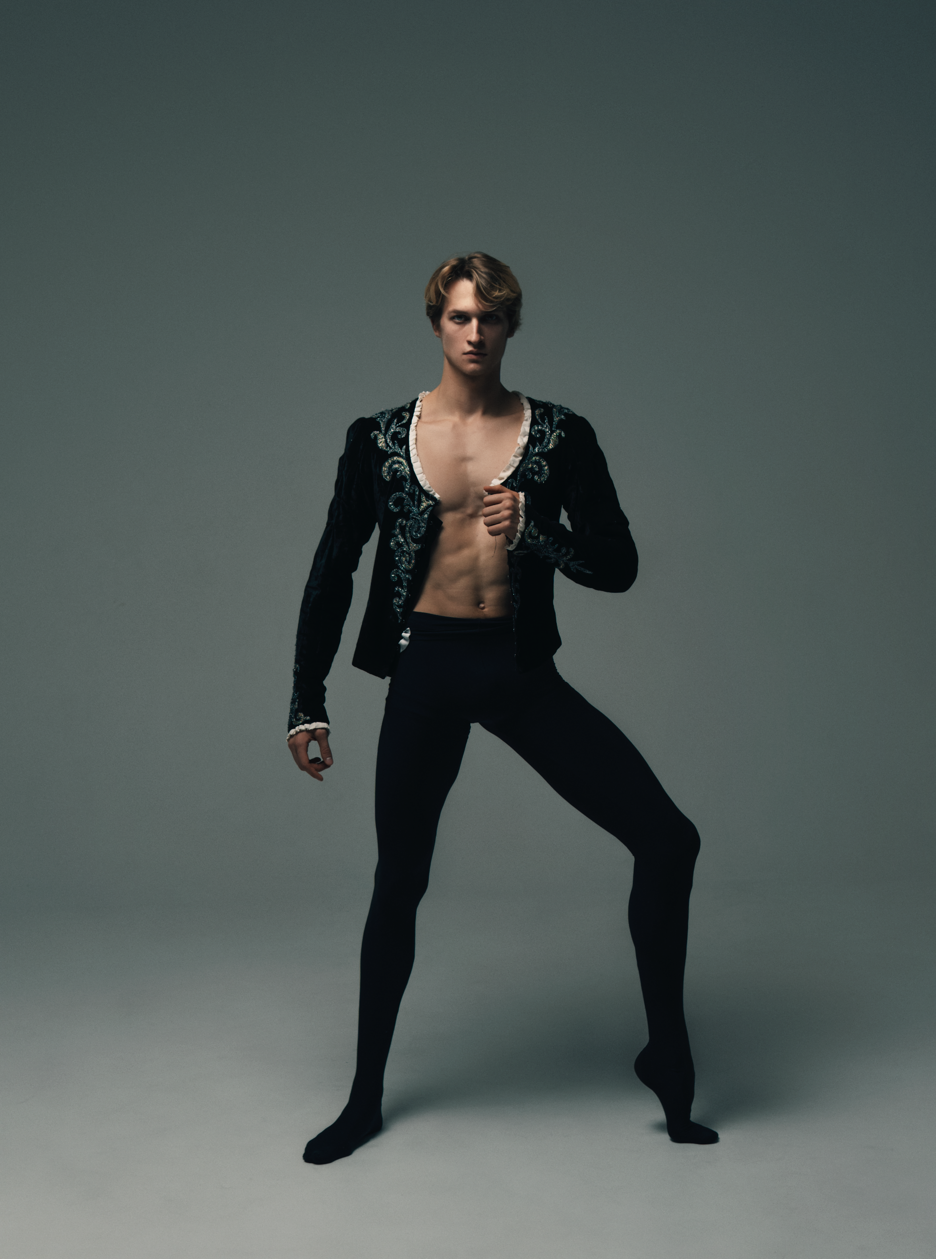 Ballet dancer in black outfit, ornate jacket, arms raised, poised pose. Gray backdrop.