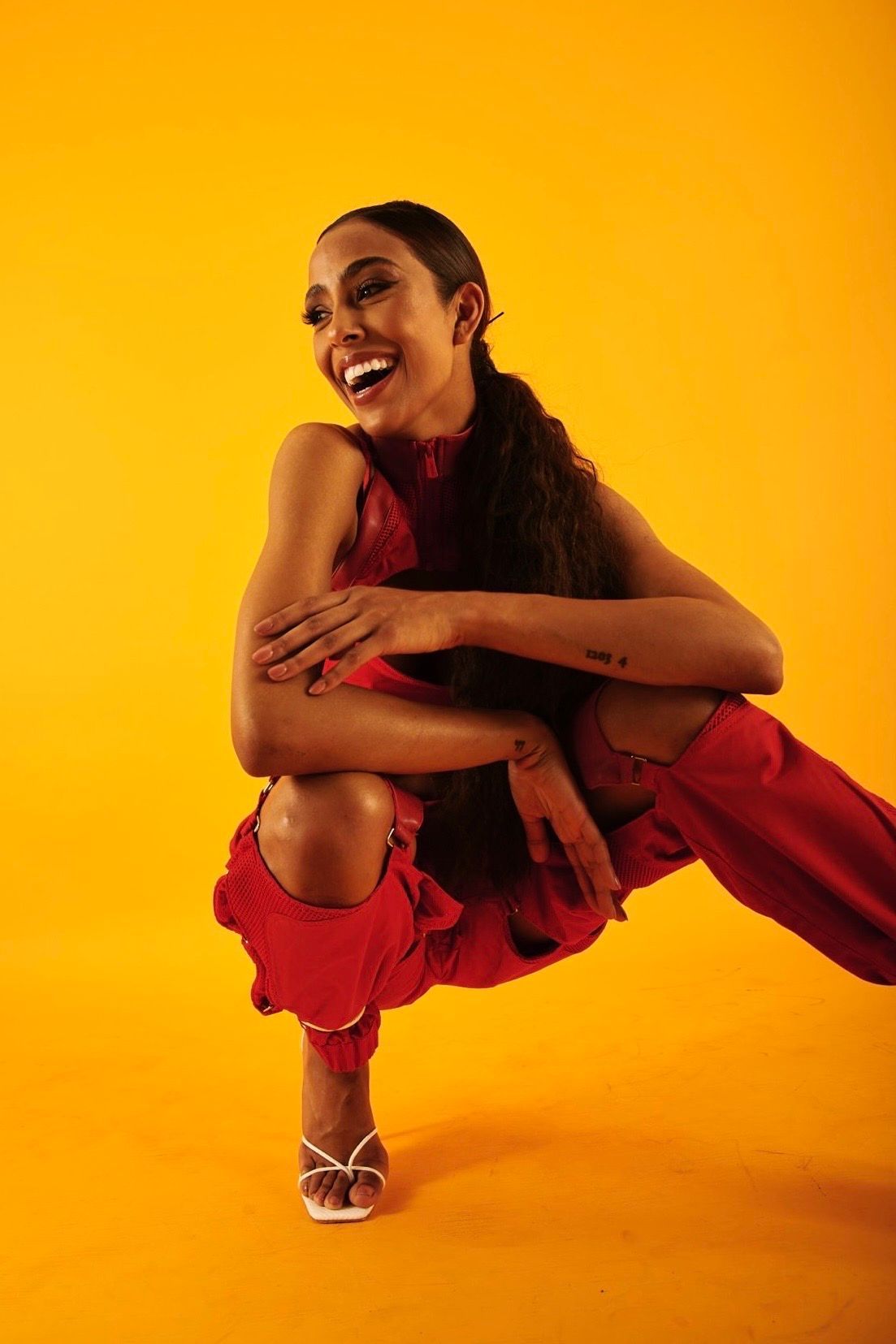 Woman in red outfit smiles, squats. Yellow backdrop.