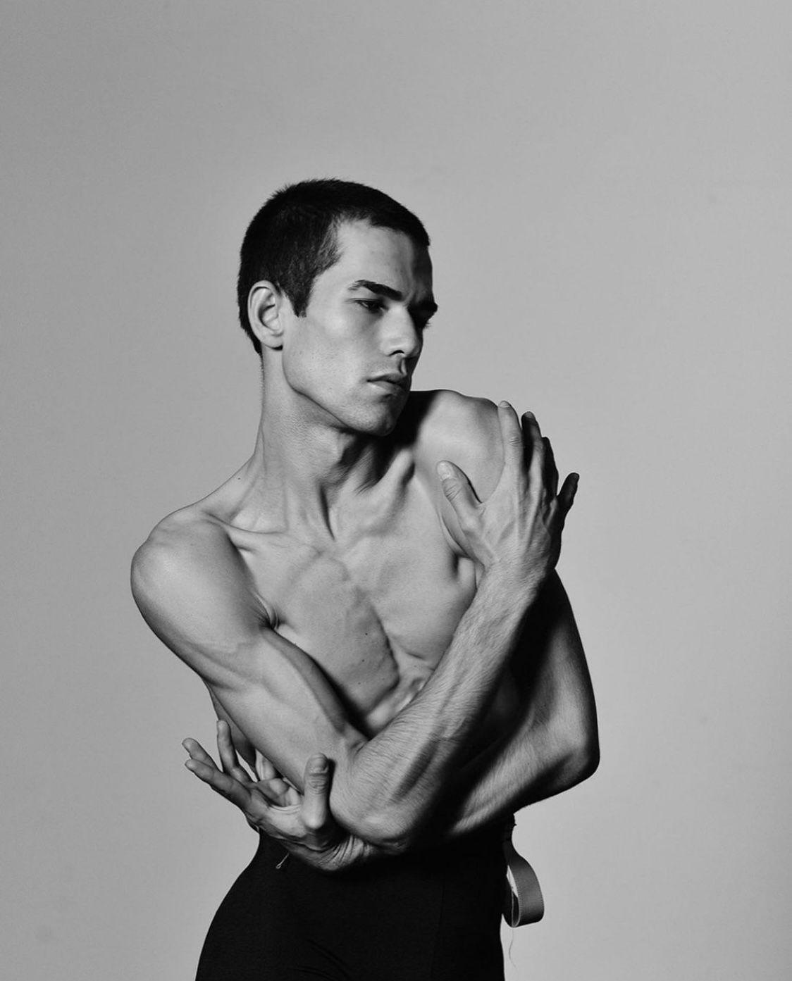 Male dancer in a graceful pose, arms crossed, torso bare, gazing to the side. Black and white.