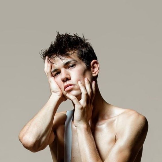 Man, shirtless, with a hand on his head and the other at his chest, looking down, pale background.