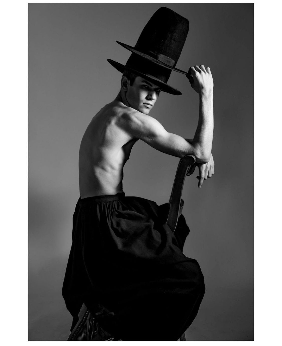 Man wearing a wide-brimmed hat, leaning on a dark object, torso exposed, looking back over shoulder.