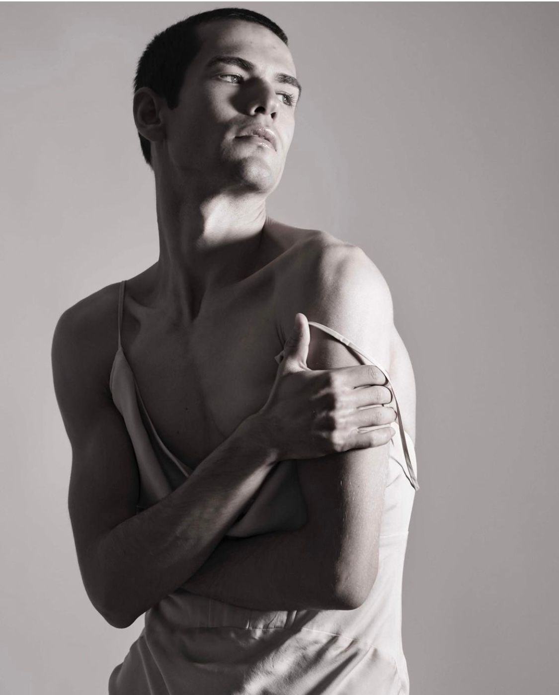 Man partially removing a light-colored tank top, looking upwards, arms crossed.