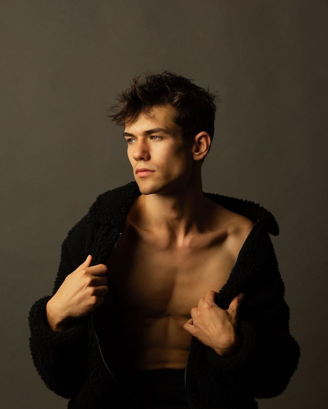 Man with open black jacket, looking to the side, against a gray background.