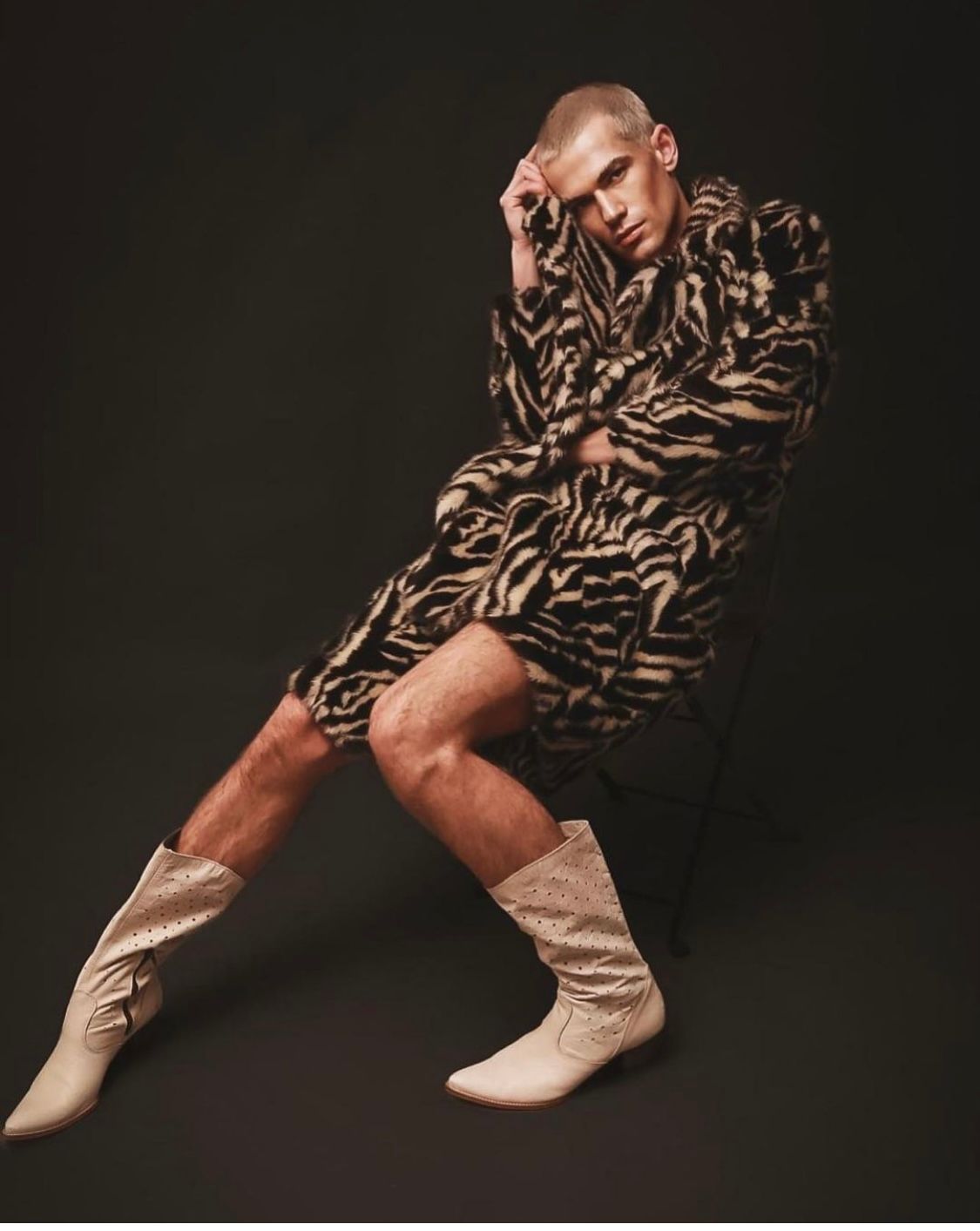 Man in zebra print coat and cowboy boots, seated with hand on head, posing against a dark background.