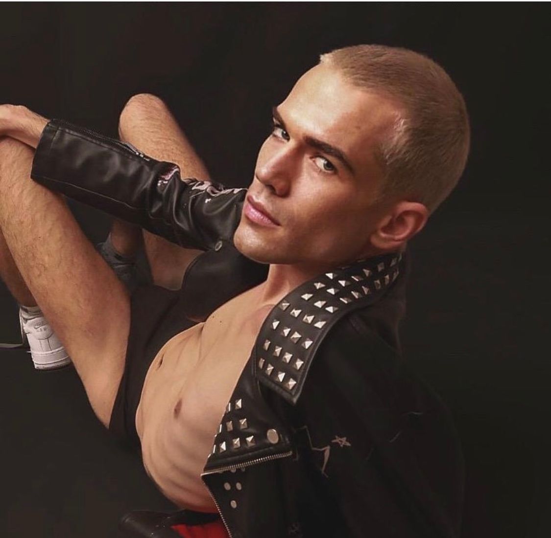 Man in studded jacket and shorts reclines, looking at the camera. Black background.