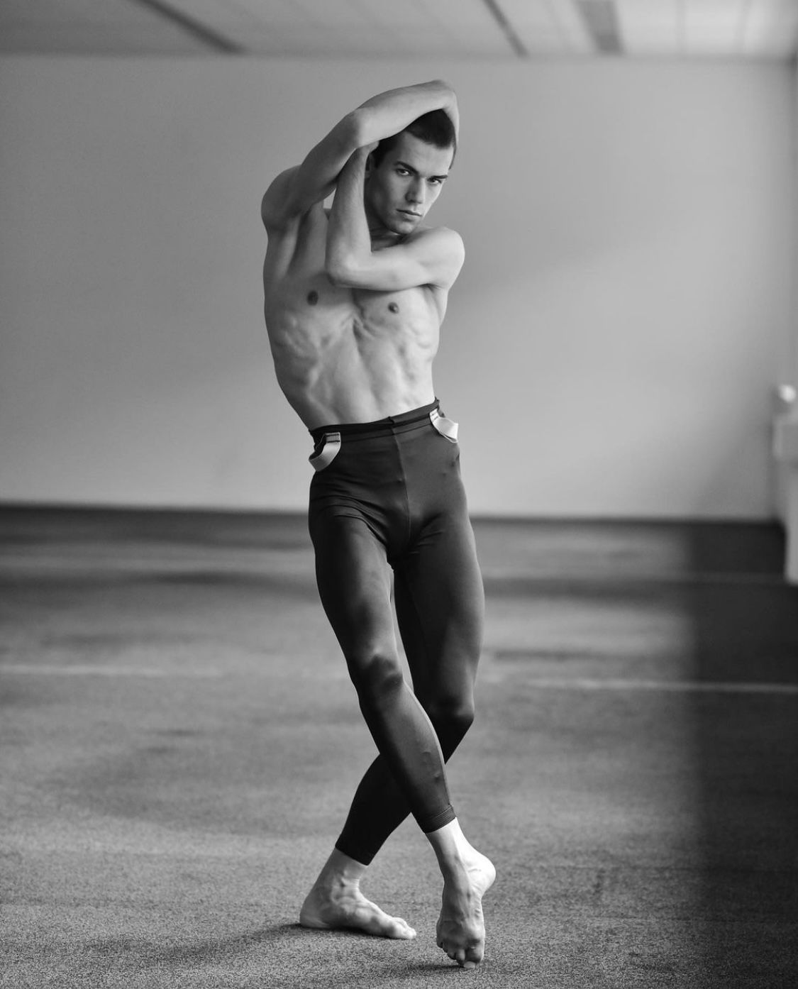Ballet dancer in a pose, arms overhead, legs crossed, wearing dark pants and bare feet.