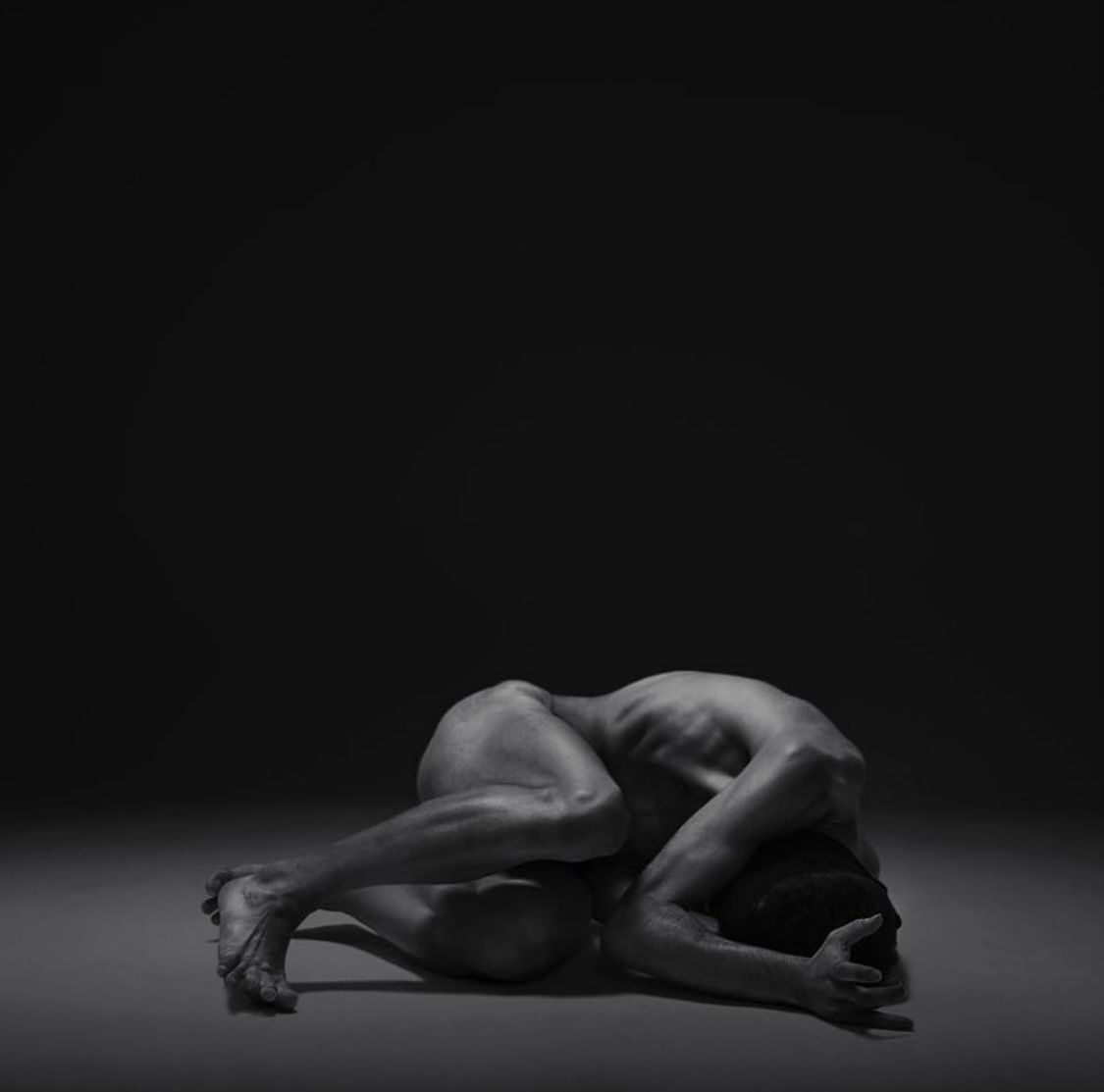 A person curled on the ground in a dark space, back arched, head down, arms over head.