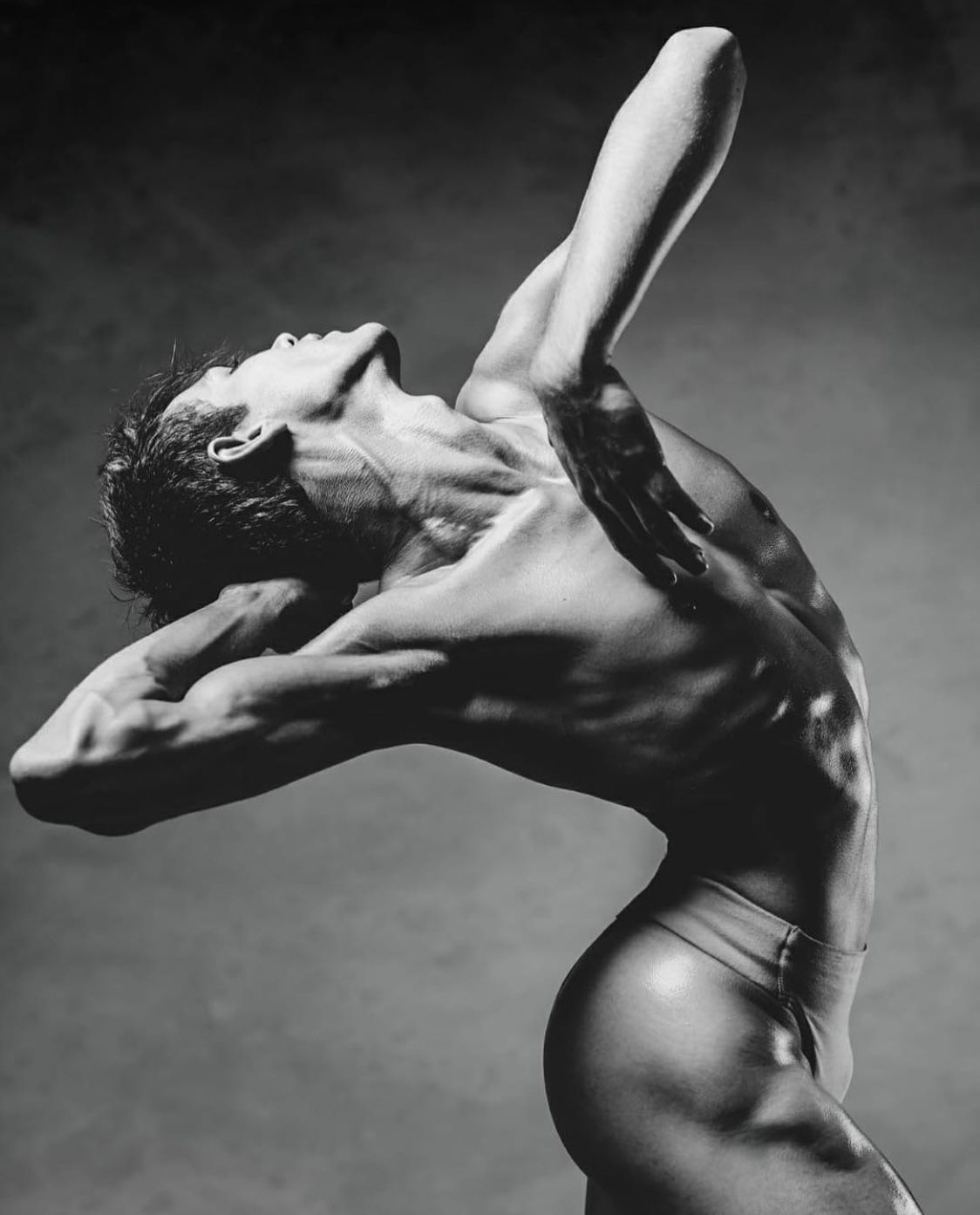 A dancer in a dramatic pose, arched back with arms raised overhead, in grayscale.