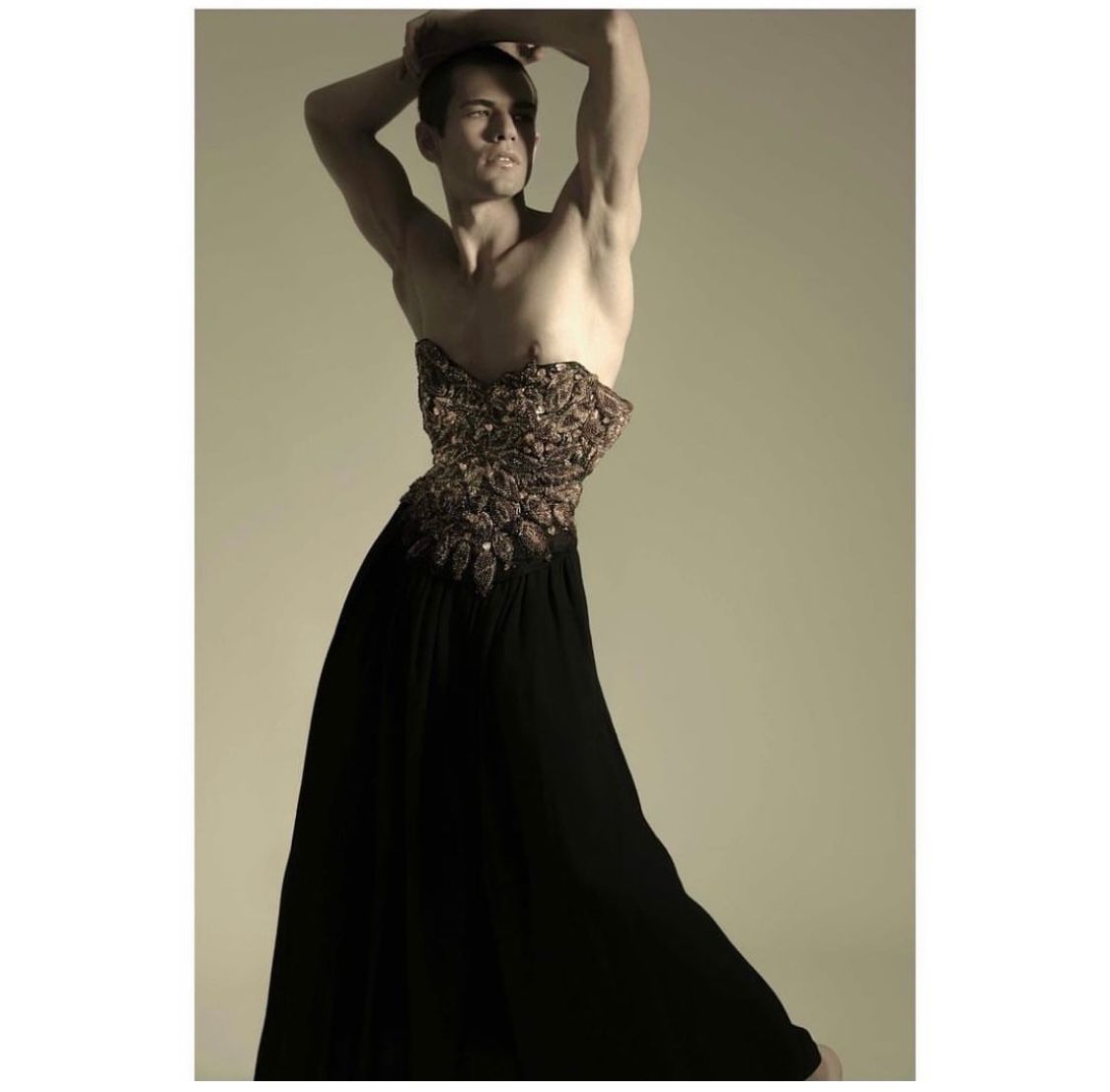 Man in strapless gown poses with arms raised; sepia tones.