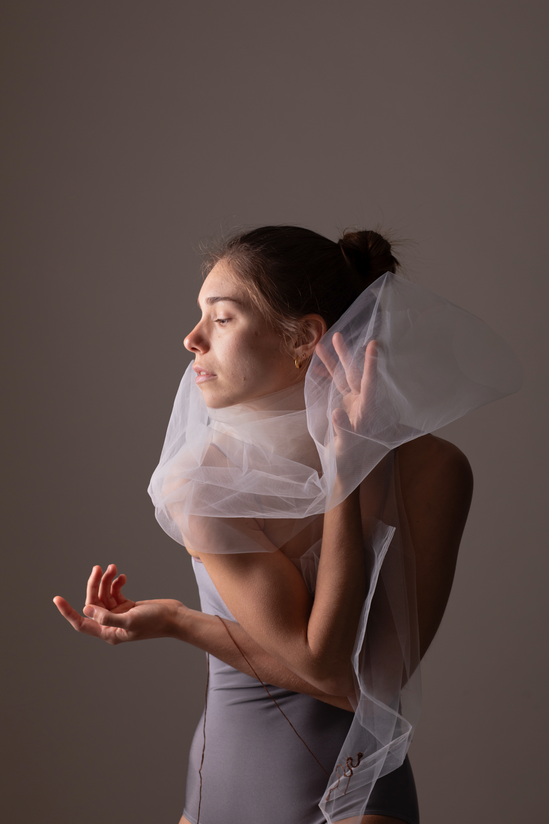 Woman in a gray leotard with a sheer white scarf, eyes closed, hands raised, against a neutral background.