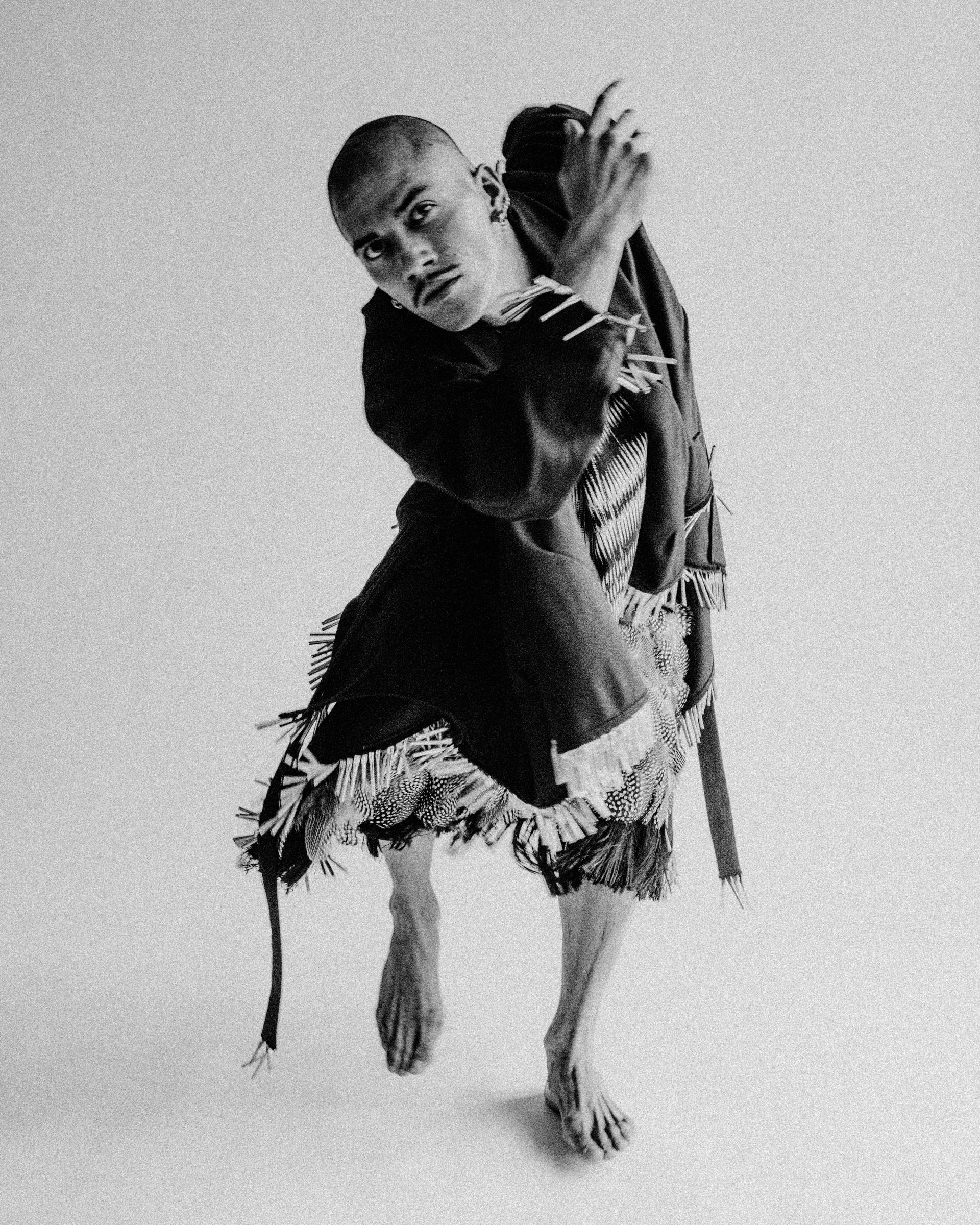Person in fringed garment, arms crossed, feet flexed, poses in front of a white background.