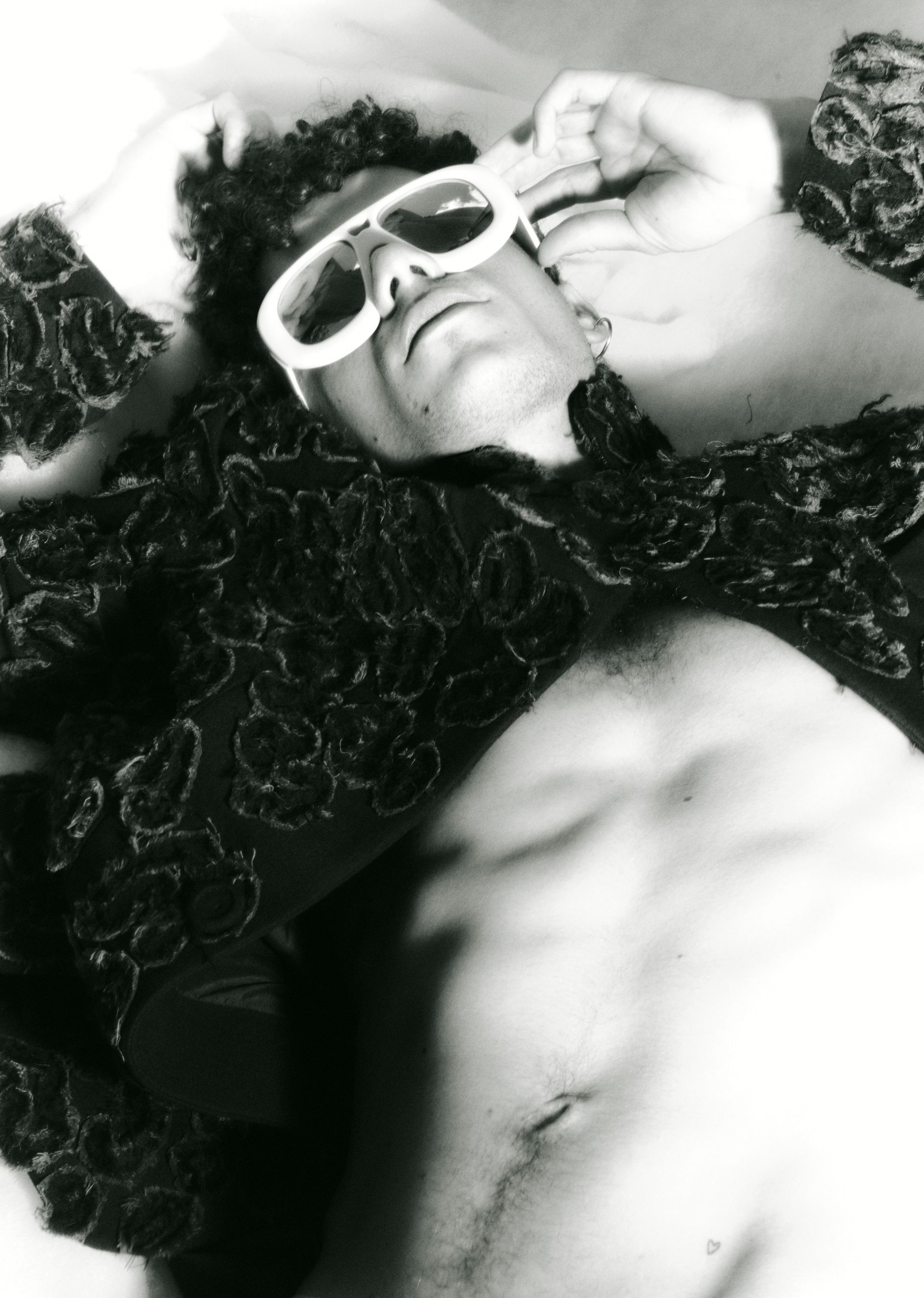 Man wearing sunglasses, lying down with open shirt. Black jacket with textured details.