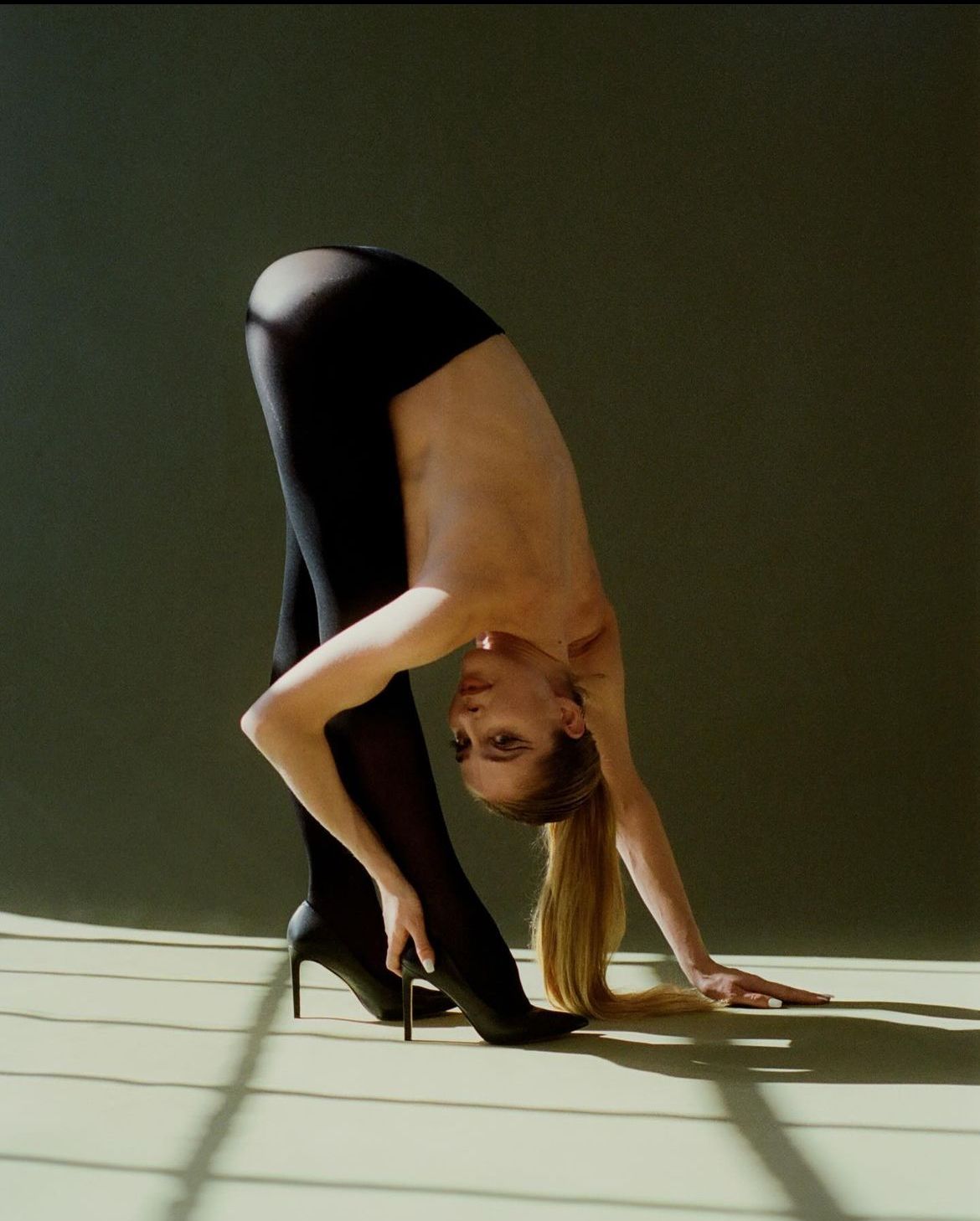 Woman in black tights and heels bending over, torso bare, touching floor.