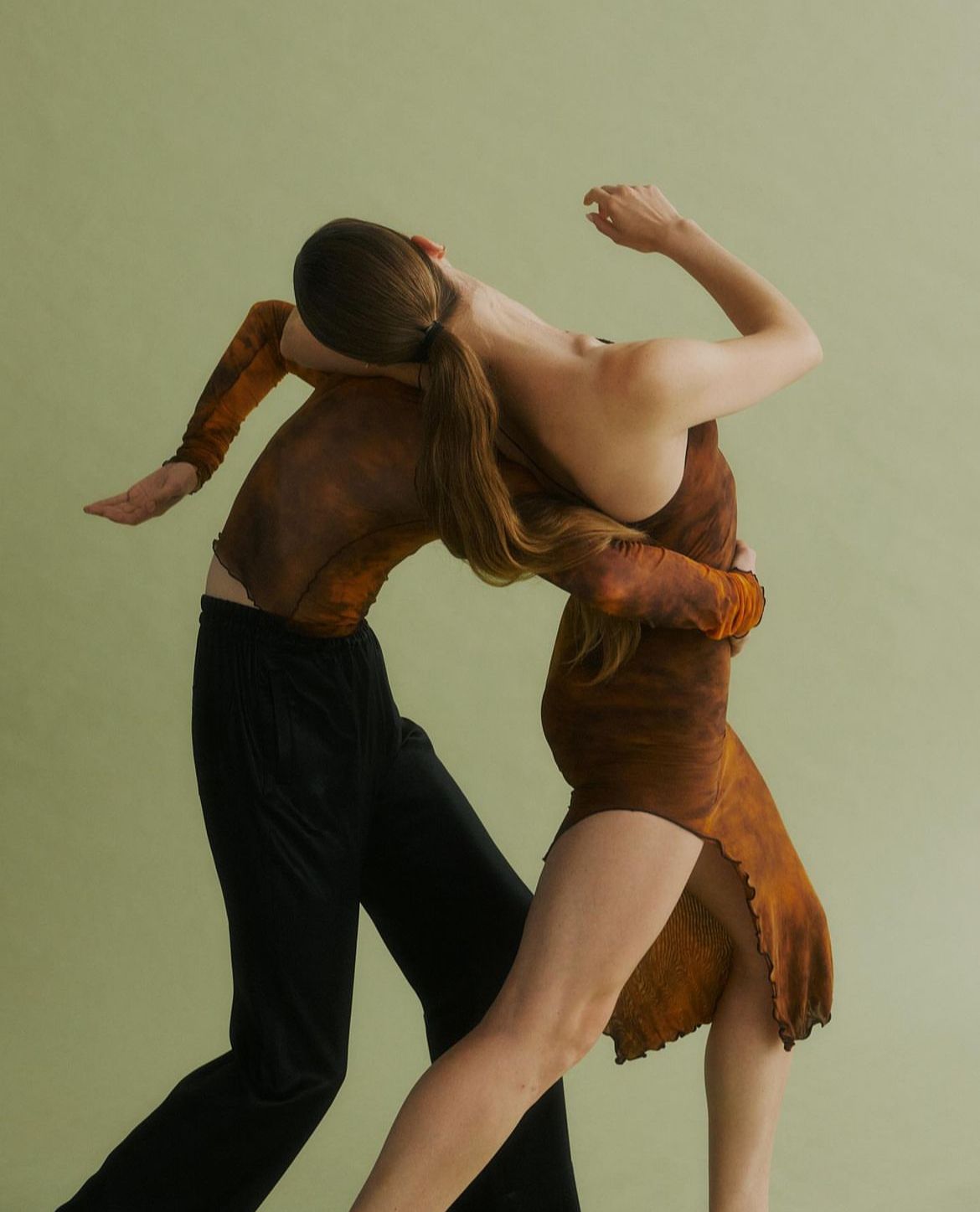 Two people in brown and black outfits embracing, arms raised, against a green background.